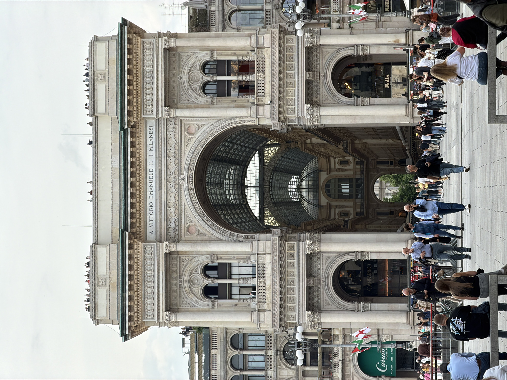 The image displays a bustling daytime scene featuring the grand architecture of the Galleria Vittorio Emanuele II in Milan, Italy. The perspective is from ground level, looking up at the ornate, multi-story structure that defines this iconic public space.
The building's facade is richly detailed with classical elements, including prominent arches, columns, intricate carvings, and patterned cornices. The distinctive glass and iron barrel-vaulted roof of the Galleria is visible through the main arched entrance and reflected in some of the upper-story windows, showcasing its impressive interior.
Numerous people populate the scene. On the paved ground level, individuals and small groups walk, stand, and gather, with some seated on benches. Many more people can be seen through the open arches, suggesting the Galleria's interior is also lively. A noticeable group of individuals is observing the scene from an upper balcony or rooftop section of the building.
Visible commercial signs include "Motta" (appearing twice, likely for a cafe or restaurant) and "CAMPARINO." Another sign, partially visible, advertises "Curran GIOIELLERIE MILANO ASTI SANA," indicating a jewelry store. Italian flags are displayed near one of the establishments. The sky is overcast, suggesting a cloudy but bright daytime. The overall impression is one of a busy urban landmark, with people engaging in everyday activities like strolling and sightseeing.