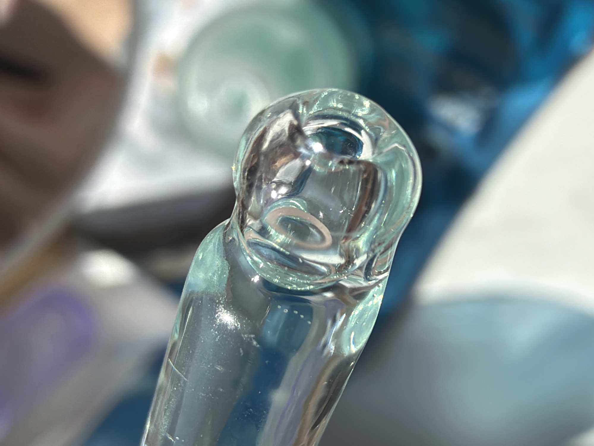 A macro shot shows the rounded top of a clear glass object, possibly a pipette or similar laboratory instrument. The glass exhibits slight bluish-green hues and contains internal reflections and refractions of light. The background features blurred, out-of-focus elements in shades of blue, white, and gray. No discernible details suggest the location is Saint Petersburg, Russia. The image does not contain people.