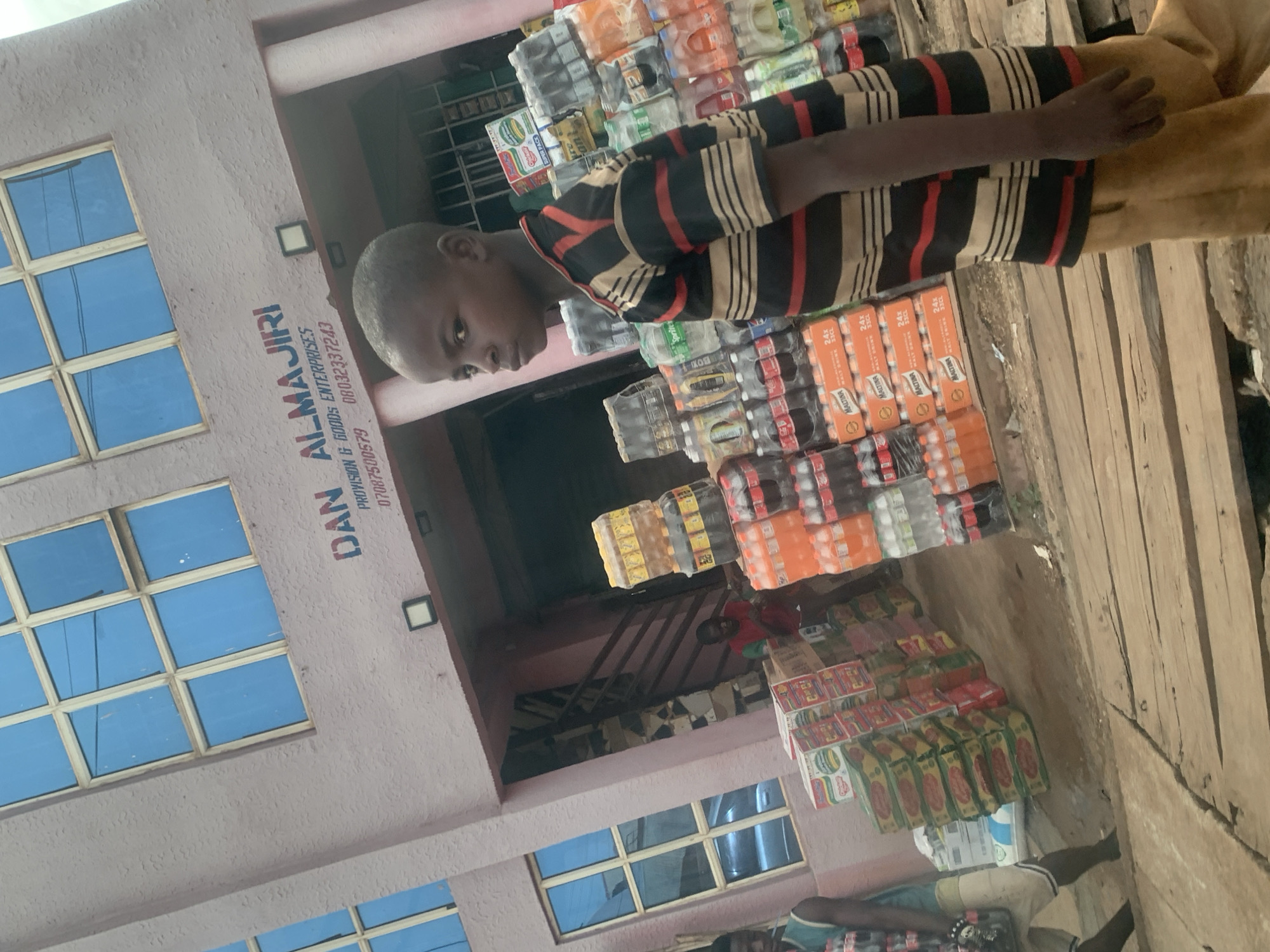 Here is a detailed description of the image:
**Setting:** The image appears to be taken at a small retail shop in Kafanchan B, Nigeria. The shop, identified by the signage "DAN ALMAJIRI," sells provisions and general goods.
**People:**
* A young boy wearing a striped shirt stands near the shop's merchandise.
* Partial views of other individuals are present near the bottom of the image.
**Objects and Merchandise:**
* Stacks of bottled drinks, including Maltina, are prominently displayed in front of the shop.
* Other food items, possibly noodles or similar products, are stacked towards the lower part of the frame.
* The shopfront features windows with a grid pattern.
* The exterior is painted in light pink.
**Actions and Interactions:** The boy's presence indicates a potential transaction or his involvement with the shop. The overall scene suggests a typical small business environment in a residential area.
**Environment:** The ground in front of the shop is wooden planks.