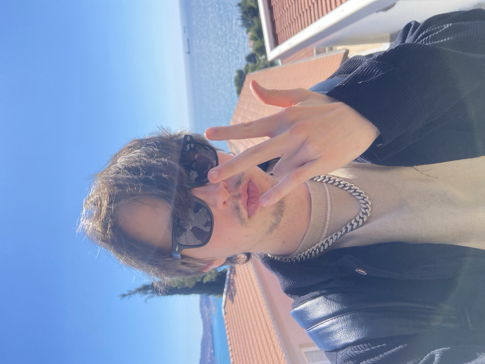 A person, likely male, is posing for a selfie outdoors on a sunny day. He has brown hair, a stubble beard, and is wearing sunglasses. He is making a peace sign with his right hand. He is also wearing a beige sweater, a black jacket, and a silver chain. In the background, there is a clear blue sky and a body of water, likely the Adriatic Sea. The presence of red tile roofs and greenery suggests the location is likely in a coastal residential area. Based on the architectural style and vegetation, the location is likely Šušanj, Montenegro. A ship is visible on the sea.