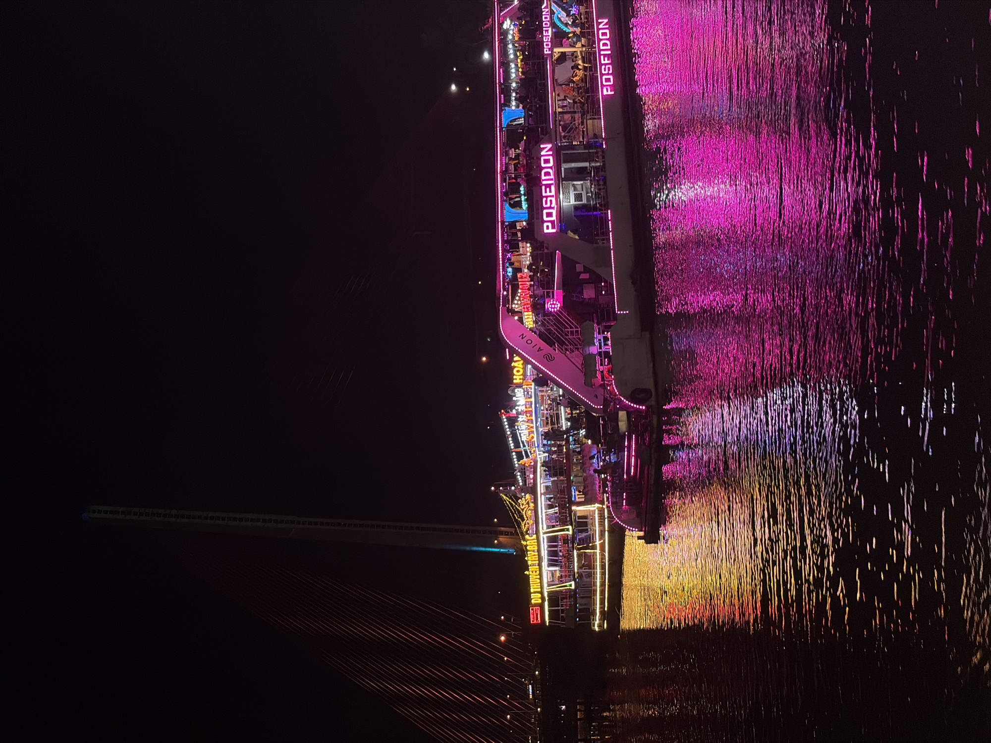 This night photograph from Đà Nẵng, Vietnam, showcases a vibrant scene dominated by a brightly illuminated cruise boat on a dark body of water, likely the Han River. The boat is adorned with numerous neon and string lights in striking colors, primarily pink, purple, and yellow.
Prominently displayed on the side of the boat are several neon signs reading "POSEIDON." Another sign appears to spell "FOSFIDON," possibly an alternative branding or a variation. Other partially visible text includes "HOÀNG YẾN," "DU THUYỀN" (Vietnamese for "cruise ship"), and "AION," suggesting different sections or operators on the vessel. Silhouettes of people can be seen on the multiple decks of the boat, indicating a lively activity, likely a night cruise or dinner.
The boat's intense lights create dazzling, elongated reflections on the water's surface, forming shimmering streaks of pink, purple, yellow, and red that ripple with the water's movement. These reflections fill the right side of the frame, adding depth and color to the dark environment.
In the background, to the left, a large suspension bridge is visible. Its structure is subtly outlined with lights, and red suspension cables are discernable against the dark sky. The overall scene conveys a festive and lively atmosphere typical of an evening attraction in a bustling city. The time of day is clearly night, with the darkness of the sky and surroundings highlighting the artificial illumination.
