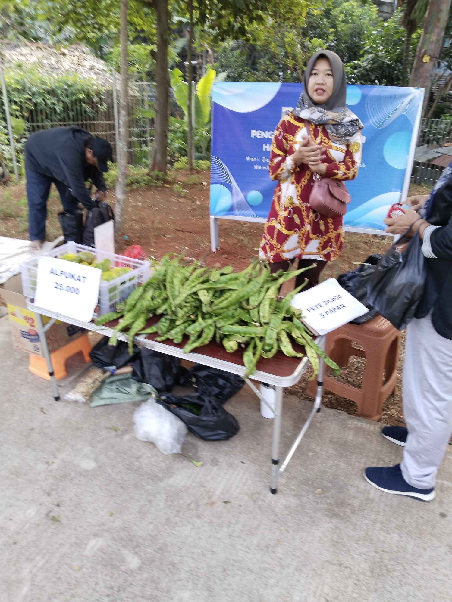 A medium shot captures an outdoor market scene where a vendor, a woman wearing a hijab and a patterned dress, stands behind a table laden with produce. To her left, a crate holds avocados with a price tag reading "ALPUKAT 25.000." A pile of long, green beans, identified by a sign as "PETE 20.000 5 PAPAN," is spread across the table. In the background, a man in a black jacket and cap bends over, seemingly organizing items. The environment includes a blue banner with text, trees, and a dirt ground. The image appears to be taken in South Jakarta, Indonesia.