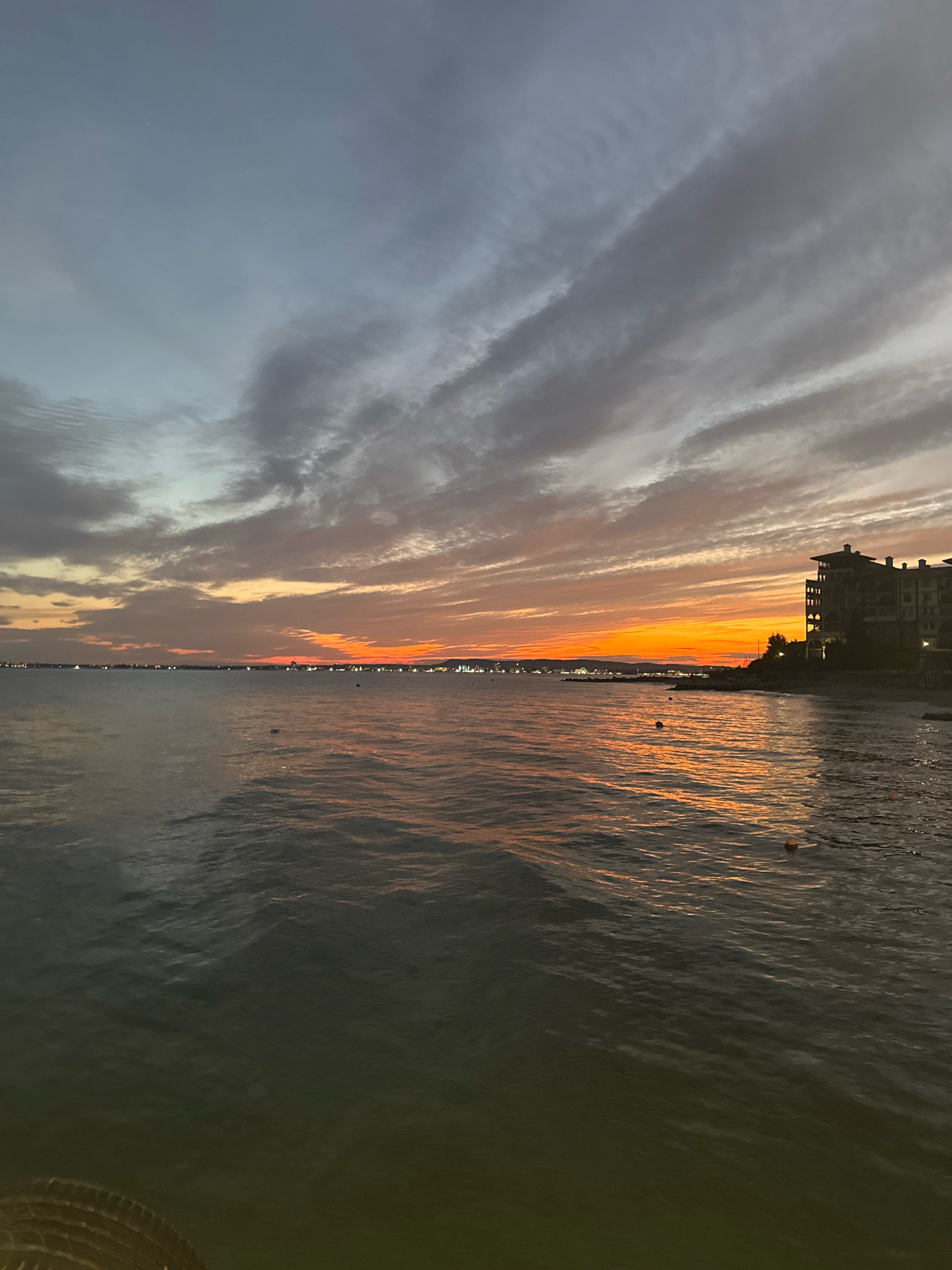The image captures a sunset over the Black Sea, viewed from the vicinity of the port in Sveti Vlas, Bulgaria. The sky is a dramatic display of colors, transitioning from a deep blue-gray at the top to vibrant oranges and reds near the horizon. Wispy, elongated clouds stretch across the sky, catching the last rays of sunlight.
In the distance, a coastline is visible, dotted with lights from buildings and possibly streetlights, suggesting a populated area. To the right of the frame, a multi-story building with a distinctive architectural style stands prominently against the sunset. Its dark silhouette is sharply defined by the bright sky.
The sea in the foreground is calm, with gentle ripples reflecting the warm hues of the sky. Several buoys are scattered on the water's surface, some marked by orange floats. The water transitions from a darker, greenish-brown in the foreground to a lighter, shimmering surface closer to the horizon, illuminated by the sunset's glow. In the lower left corner of the image, a portion of a woven object, possibly a basket or mat, is visible, suggesting the vantage point is from a waterfront structure or a beach. The overall atmosphere is serene and picturesque, typical of a coastal sunset.