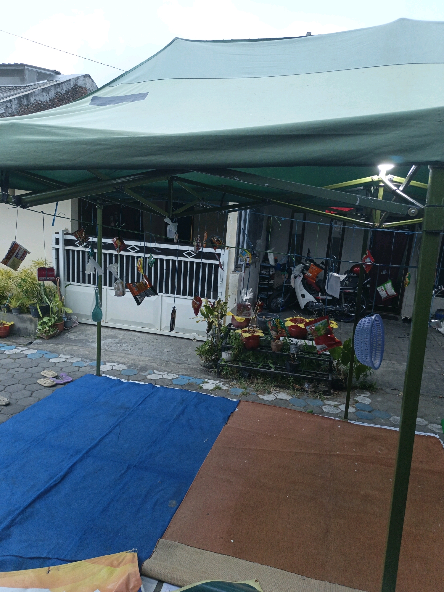 The image depicts an outdoor scene under a light green canopy tent. The ground is covered with a blue mat on the left and a brown mat on the right. In the background, a house with a white gate and fence is visible. Several motorcycles are parked in front of the house. Various items, including what appear to be empty food wrappers and plastic containers, are hanging from strings under the tent. A blue plastic basket is hanging on the right side of the frame. Potted plants are arranged on a metal rack in front of the house. The location is identified as Karangploso, Indonesia. The sky is overcast.
