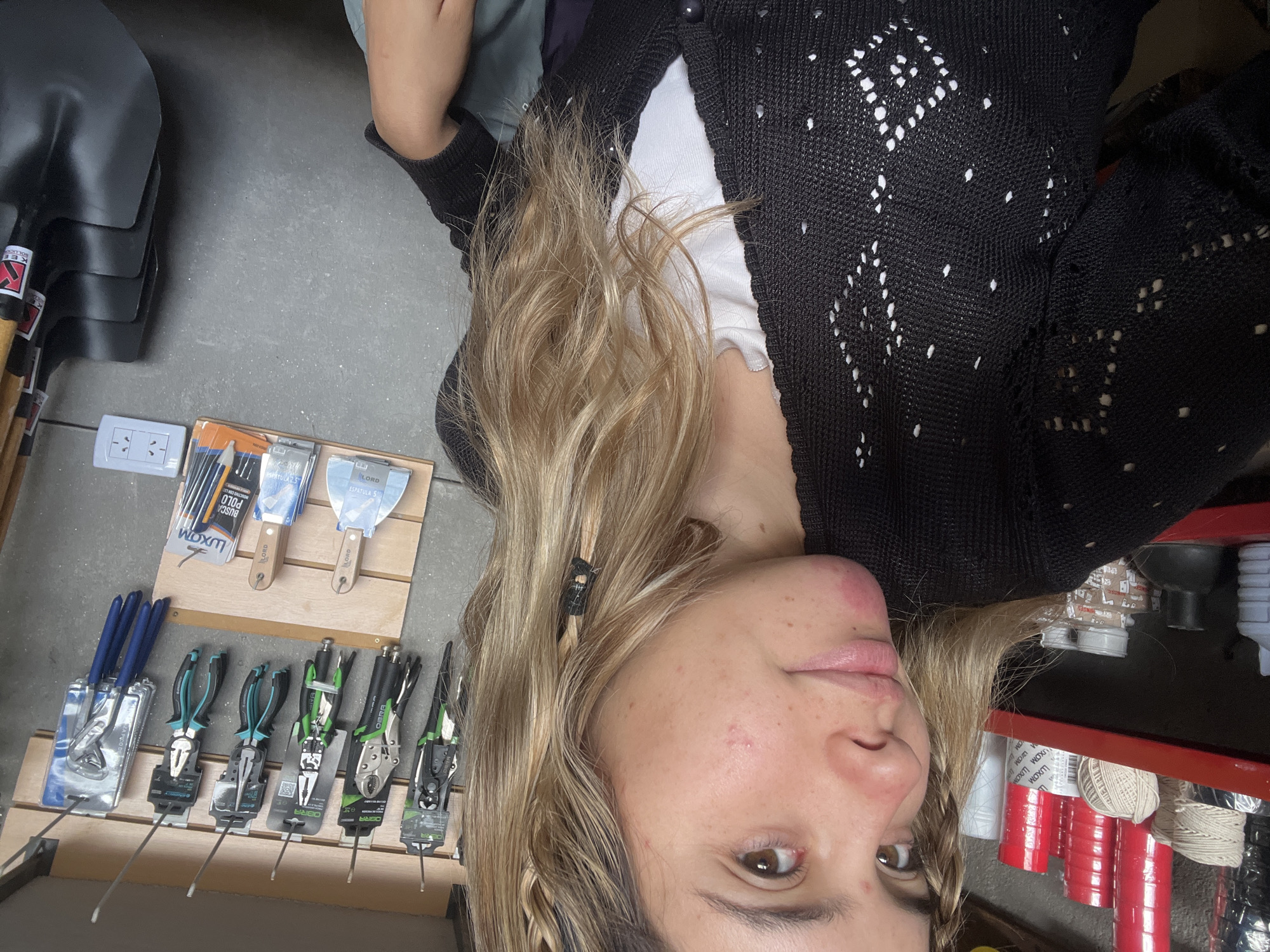 A person is facing the camera, with their head tilted slightly to the left. The individual has blonde hair, light skin, and appears to be standing in a hardware or home improvement store. To the left are tools, including pliers and spatulas, arranged on a wooden display shelf. Behind and to the right, partially visible, are items like jars, cans, and spools of twine on a red shelf. The person is wearing a black cardigan with a white shirt underneath. The photo seems to have been taken from a close perspective. The geographic location, Sumalao, Argentina, is not evident from the image.
