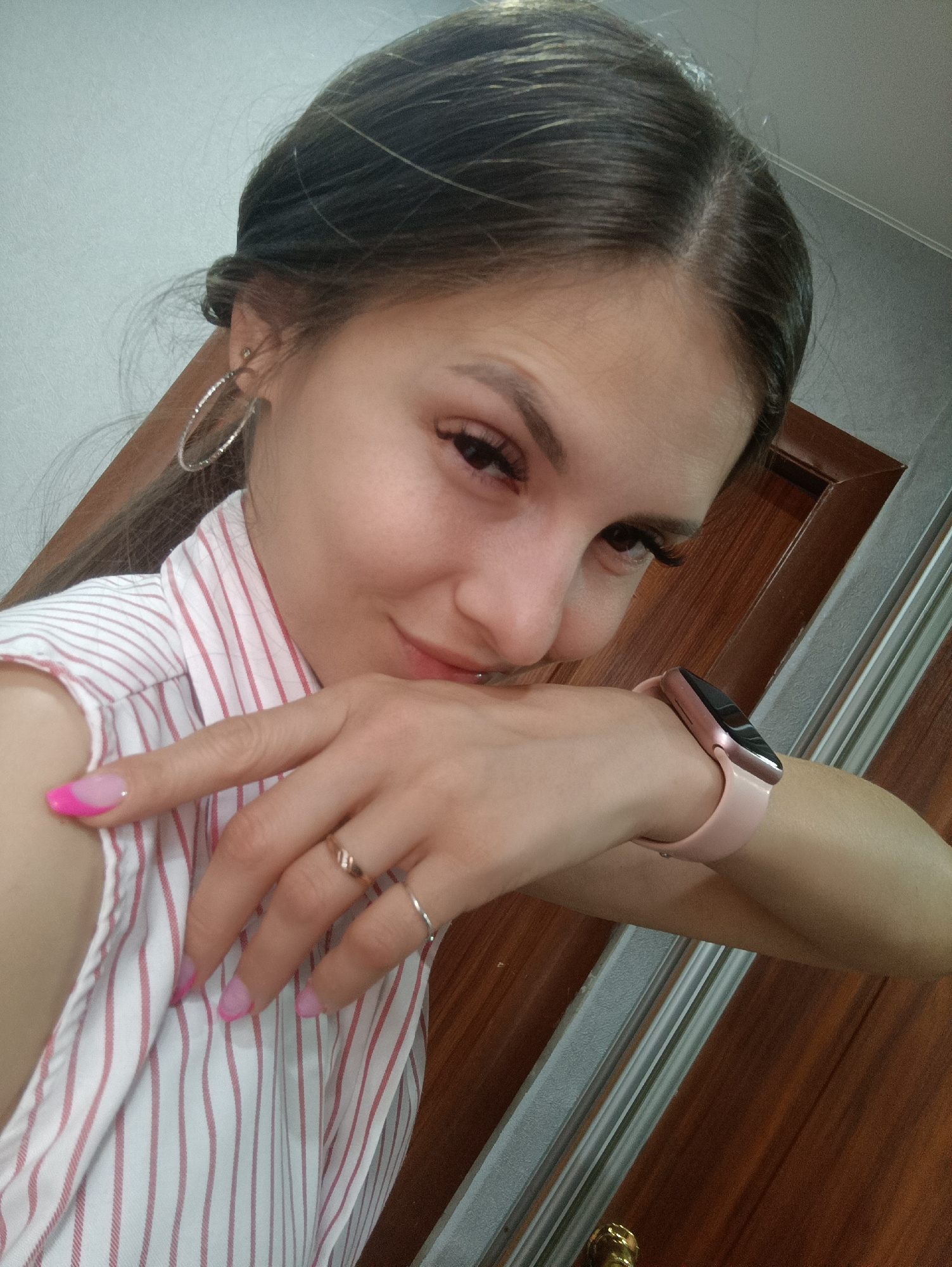 A young woman is photographed from the chest up, looking down and to the right with a slight smile. She has dark hair pulled back, large hoop earrings, and wears a white and red striped shirt. Her left hand is raised to her face, with painted nails, and she is wearing rings. A pink smartwatch is visible on her right wrist. The background appears to be indoors with wooden doors and a white wall.