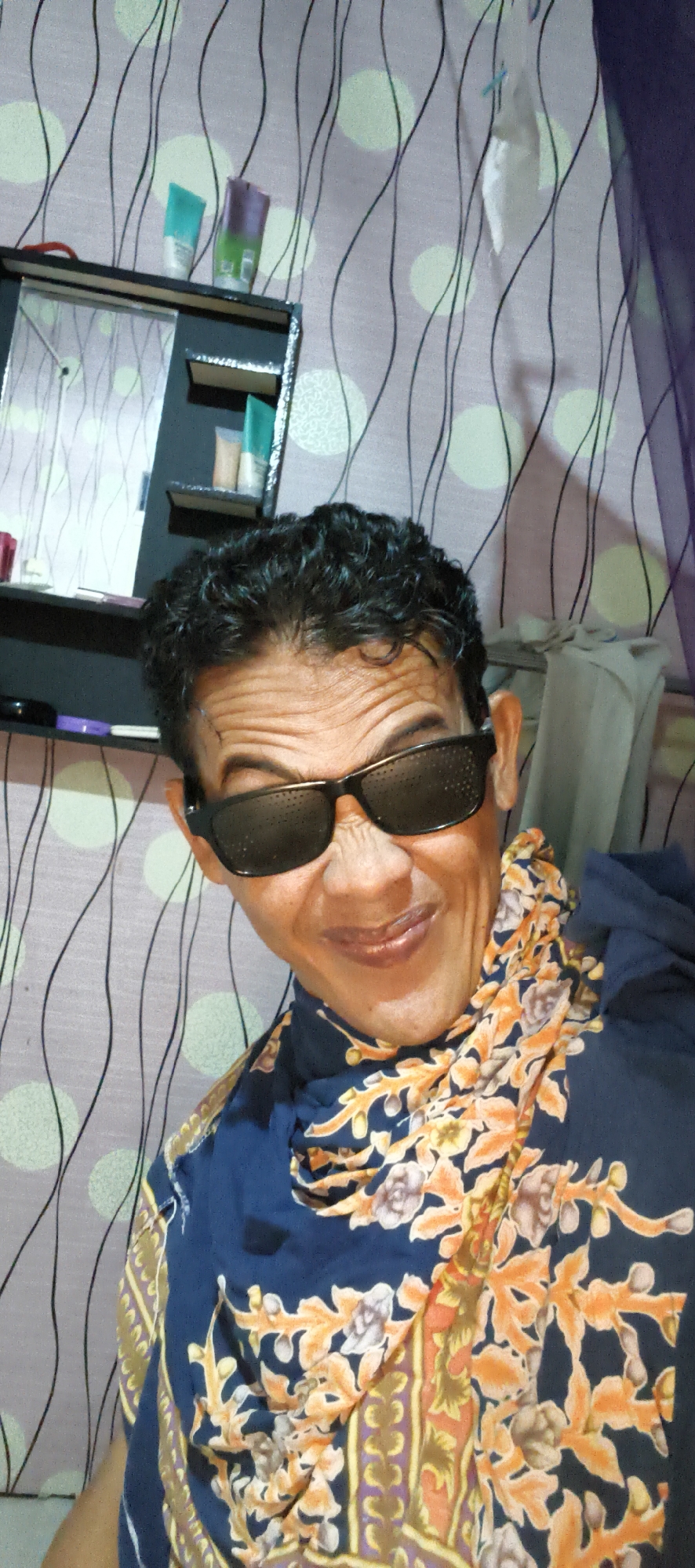 A person is taking a selfie indoors, possibly in a bedroom or bathroom, given the presence of a shelf with toiletries and a mirror. The person is wearing dark sunglasses and a patterned scarf or wrap over a dark blue shirt. The background appears to be a wall with a patterned wallpaper featuring wavy lines and circles. The lighting is dim, and the overall impression is a casual, personal moment captured on camera. There is no discernible indication of the location being specifically in Muaro Jambi, Indonesia, beyond the user's prompt.