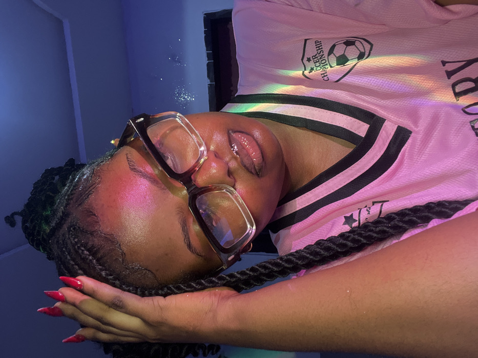 An individual, identified as a female, is depicted in a medium close-up portrait within an indoor setting in Ipetumodu, Nigeria. The subject possesses dark skin, is wearing large, clear-framed glasses with dark temples, and has braided dark hair styled upwards. Her right hand, adorned with long, pointed red acrylic nails, rests on her head near the hairline. She is wearing a pink V-neck jersey featuring black stripes on the collar and shoulders, and a "SOCCER CHAMPIONSHIP" logo with a soccer ball on the upper right chest area. A distinct rainbow light effect, along with pink, blue, and green ambient lighting, illuminates her face and upper torso, casting a prominent pink highlight on her forehead and glistening her lips. The background is indistinct, appearing as a dark blue surface.