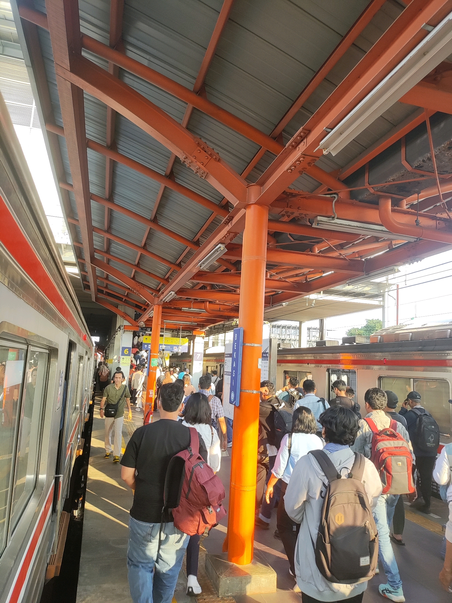 The image depicts a busy train station platform in Bekasi, Indonesia, likely during daylight hours. A train, with its silver exterior and red stripe detailing, is stopped at the platform, its doors open. A crowd of people are standing on the platform, some are boarding or alighting the train, while others are waiting. The platform is covered by an orange, exposed metal roof structure supported by orange pillars. The ground appears to be a concrete platform. Signage is visible, including a blue sign with white text that partially reads "BEKASI". People are dressed in casual clothing, and many are carrying backpacks. The scene suggests a typical commute or travel situation.