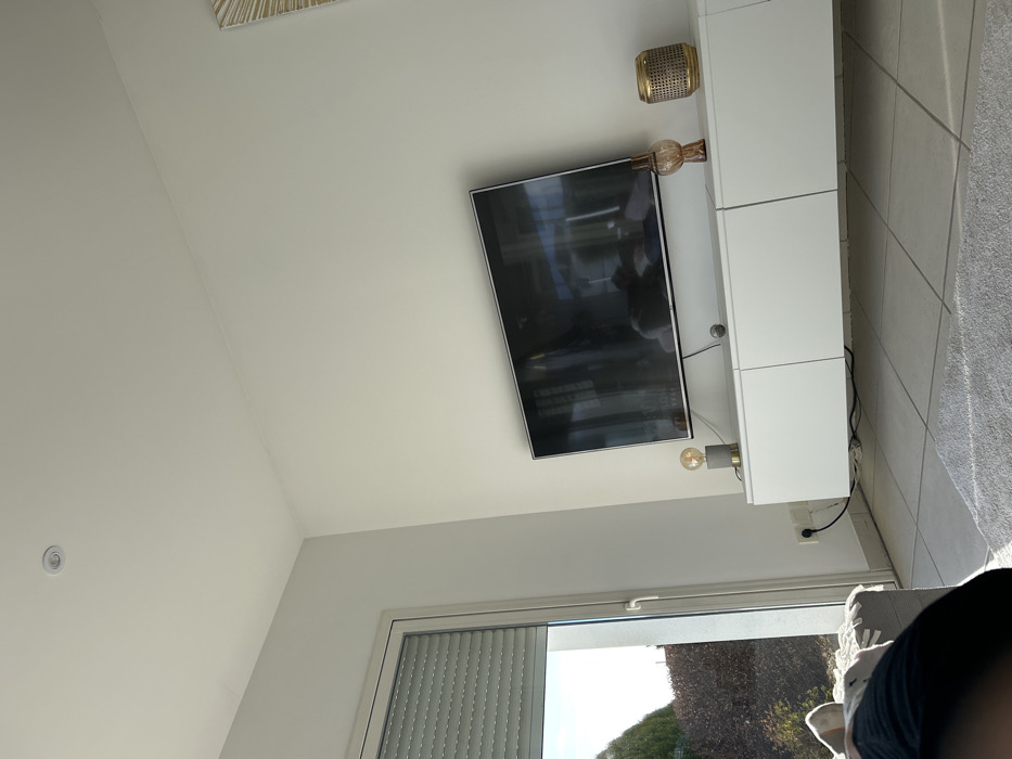 The image depicts an indoor residential space, likely a living room, located in Veauchette, France. The room features white walls and a white ceiling with a single circular recessed light fixture. A large, dark-screened flat-panel television is mounted centrally on a wall. Below the television, a white floating media console, comprising three cabinet units, is affixed to the wall.
Upon the console, from left to right, are: a decorative lamp with a grey concrete-like base, a brass-colored accent, and an exposed filament bulb; a small dark spherical object; an amber-colored glass vase with a textured surface; and a cylindrical, gold-colored woven decorative object. Reflections on the television screen show elements of the room, including what appears to be a light-colored sofa. A partial view of a decorative wall hanging with a sunburst pattern is visible on the upper right wall.
On the left side of the room, a large white-framed glass door or window is partially open or closed. A grey, horizontally slatted roller shutter is partially retracted at the top of this opening. Sunlight streams into the room through this window, illuminating the tiled floor and a light-colored, shaggy rug in the foreground. Outside the window, green foliage and brown leafless bushes are visible under a bright sky. A white electrical outlet with a black cord plugged in is situated on the wall below the window frame. In the lower-left foreground, a human leg, clothed in dark fabric and light-colored socks, rests on a light-colored textile, presumably part of a sofa or chair. No other individuals or clear actions are discernible.