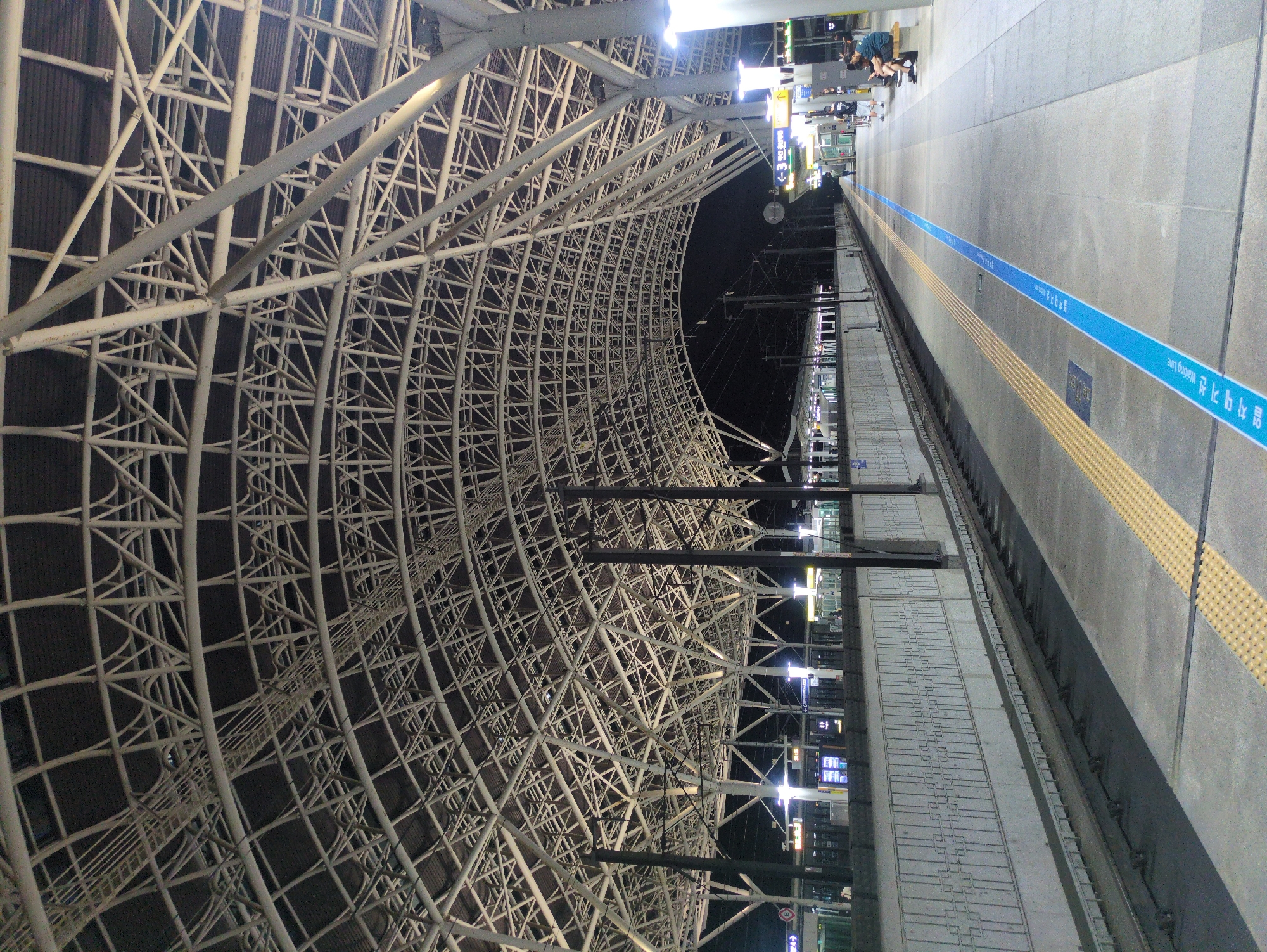 The image shows a train platform at Singyeongju Station in Gyeongju-si, South Korea, at night. The platform is illuminated by overhead lights. The distinctive architecture is a large, curved, white steel framework covering the platform. A tactile paving strip and a blue line with Korean writing run along the platform edge. People are sitting and standing on the platform in the distance.