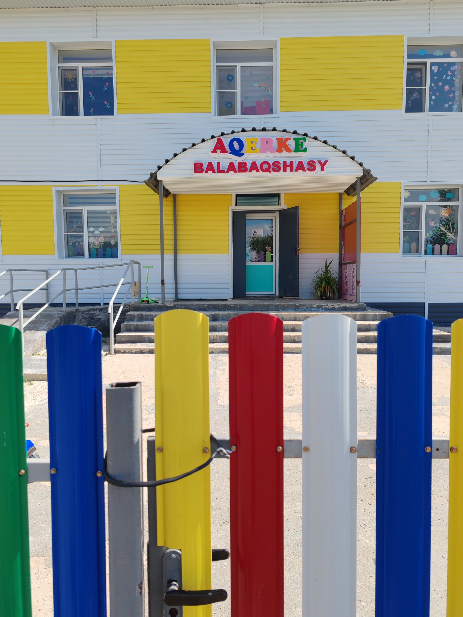 The image shows a colorful kindergarten building in the village of Akzhar, Kazakhstan. The building has yellow and white siding, with several windows decorated with various colorful images. Above the entrance, a sign reads "AQERKE BALABAQSHASY" in large, multi-colored letters. The entrance features an open door with a turquoise panel. In front of the building, a brightly colored fence made of vertical metal panels in green, blue, gray, yellow, and red separates the viewer from the scene. Stairs lead up to the entrance, and a green scooter is visible on the right side of the stairs. The environment appears to be outdoors on a sunny day, with a sandy or gravelly ground surface.