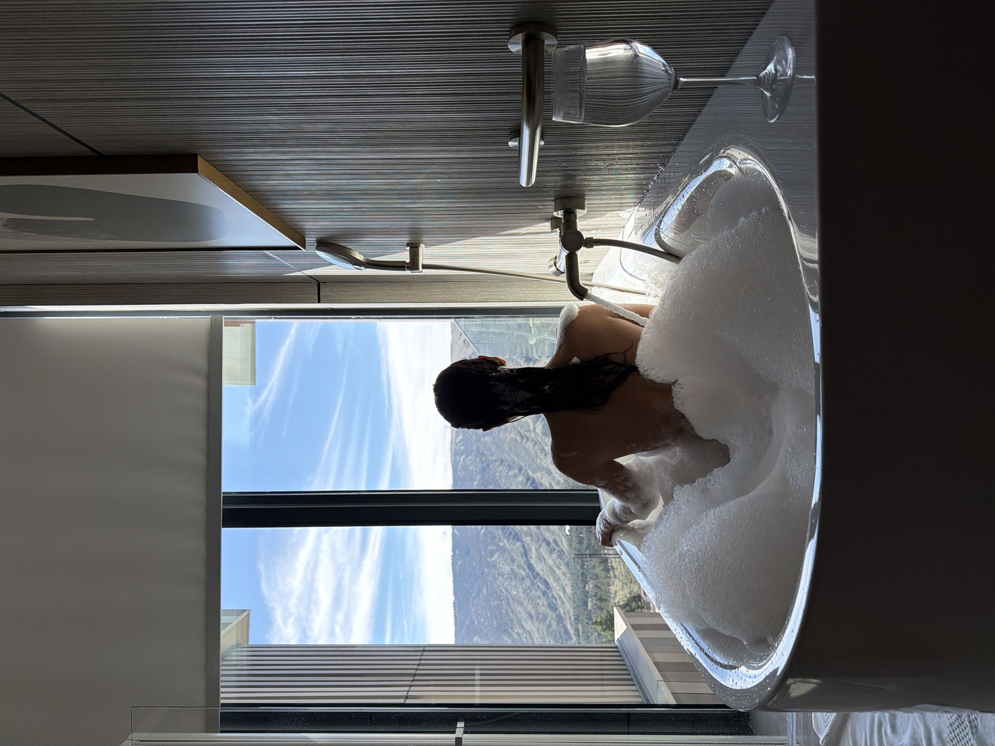 A person is in a bathtub filled with bubbles. There is a window behind the bathtub. The scene appears to be in a modern bathroom with gray tile. Outside the window is a landscape with mountains and a blue sky with wispy clouds. A wine glass is on the edge of the tub, near the faucet. The location is likely in a hotel in Tbilisi, Georgia.