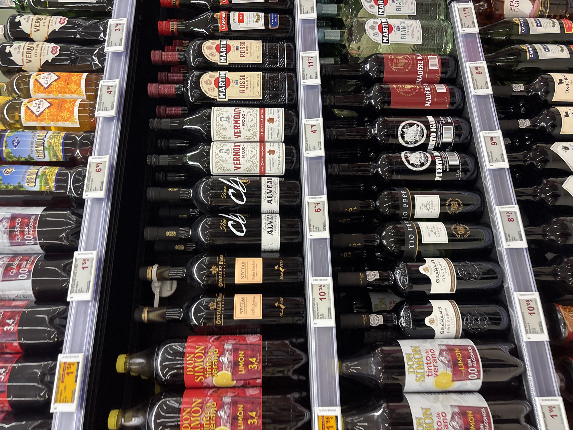 The image depicts a shelf in a Spanish supermarket in Urbanización Villa Martín, Spain, stocked with a variety of bottled alcoholic and non-alcoholic beverages. The top shelf displays larger bottles of wine and liqueurs, including brands like "Clásico," "Granado," and "La Cartuja." The middle shelf is densely packed with bottles of vermouth, sherry, and port wine, featuring prominent brands such as "Gonzalez Byass Nectar," "Conde de Cortina," "Martini Rosso," "Campane," "Don Simon Tinto de Verano," and "Graham's Fine Tawny." The bottles are primarily dark glass with colorful labels. Below, another shelf is visible, also lined with bottles, suggesting a multi-tiered display. The lighting is bright and even, typical of a retail environment. No people are present in the image, and the focus is entirely on the product display. The scene is clean and organized, indicating a well-maintained retail space.