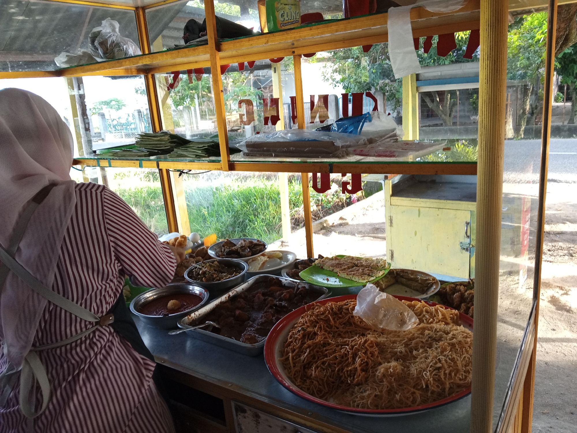 A person wearing a pink and white striped shirt and a light pink hijab stands in front of a food stall. The stall is made of wood with glass panels and is filled with various food items displayed in metal and ceramic containers. In the foreground, there is a large red and white circular dish filled with fried noodles. To the left, a metal rectangular pan holds a dark brown stew, and a circular metal bowl contains a reddish sauce with a dollop of something on top. Behind these, several other dishes of food are visible, including what appears to be fried meat and vegetables. On the shelves above, there are packaged goods and more food items. The stall is situated outdoors, with a blurred background of greenery, trees, and a road, suggesting an urban or roadside setting in Uruekanoe, Indonesia. The lighting indicates it is daytime, with sunlight casting shadows.