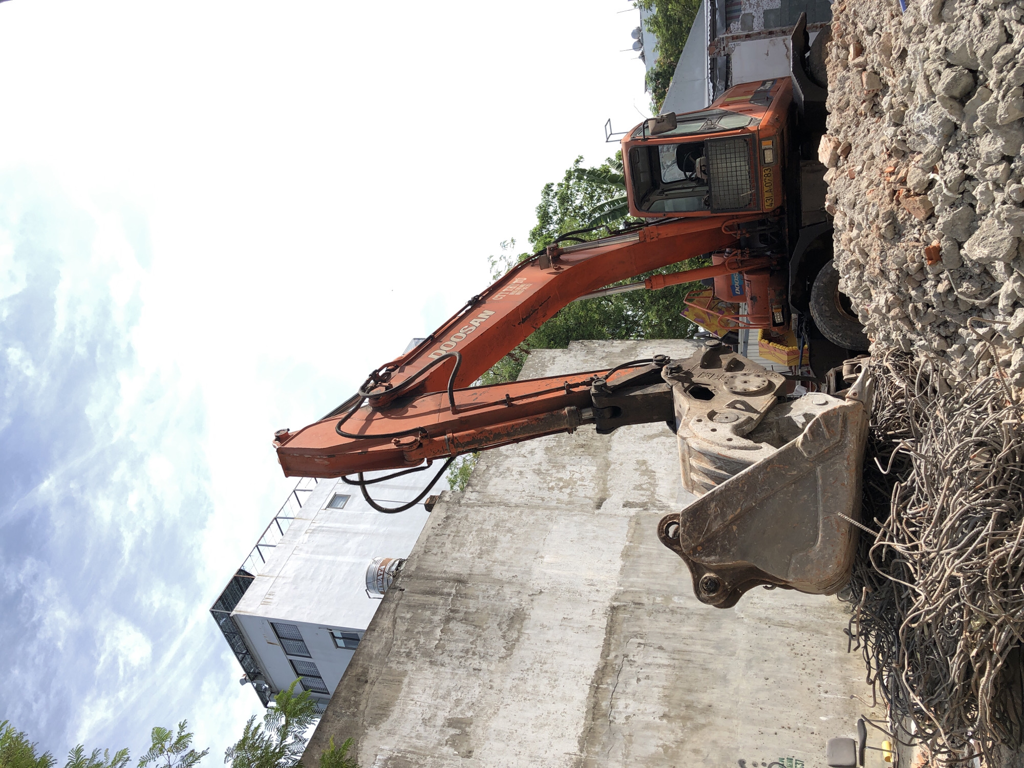 The image depicts a construction site in Da Nang, Vietnam, on a clear, sunny day. In the foreground, a pile of tangled rebar and crushed concrete is visible, with an excavator's large metal bucket resting on top of the rebar. The excavator, an orange Doosan model, is positioned in the center of the frame, its arm extended and bucket tilted. Behind the excavator, a weathered concrete wall dominates the mid-ground, showing signs of age and cracking. Above the wall, a white multi-story building with multiple windows and a rooftop structure can be seen. Green trees are present on the left and right sides of the frame, bordering the construction area. The sky is a bright blue with scattered white clouds. The overall scene conveys a sense of demolition or ongoing construction work in an urban setting.