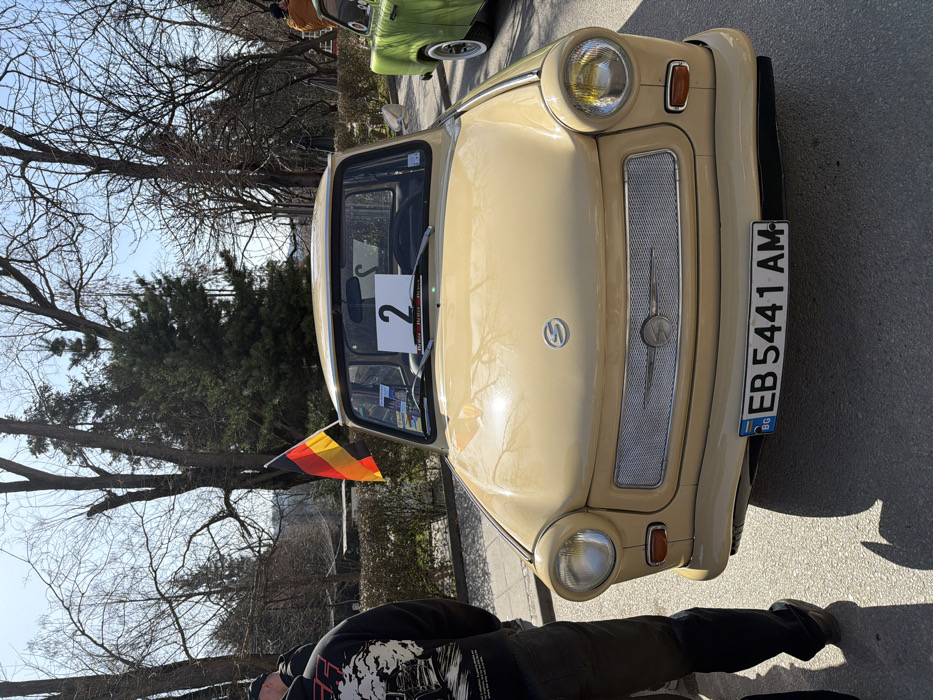 A beige Trabant car is prominently featured in a front-quarter view, parked on a paved surface under clear, sunny skies in Veliko Tarnovo, Bulgaria. The vehicle displays a Bulgarian license plate 'BG EB 5441 AM'. A small German flag (black, red, yellow vertical stripes) is affixed to the passenger side front fender. A white rectangular sign with the black numeral '2' is positioned on the dashboard. To the left, the lower body of an individual wearing dark pants and a black graphic hooded sweatshirt is partially visible. The front section of a green vintage car with white-walled tires is partially visible on the right. The background comprises a public park environment with a mix of deciduous trees (some bare) and evergreen trees. Shadows on the pavement indicate bright daylight. Distant figures are faintly discernible within the background landscape. The scene depicts a vintage car display.