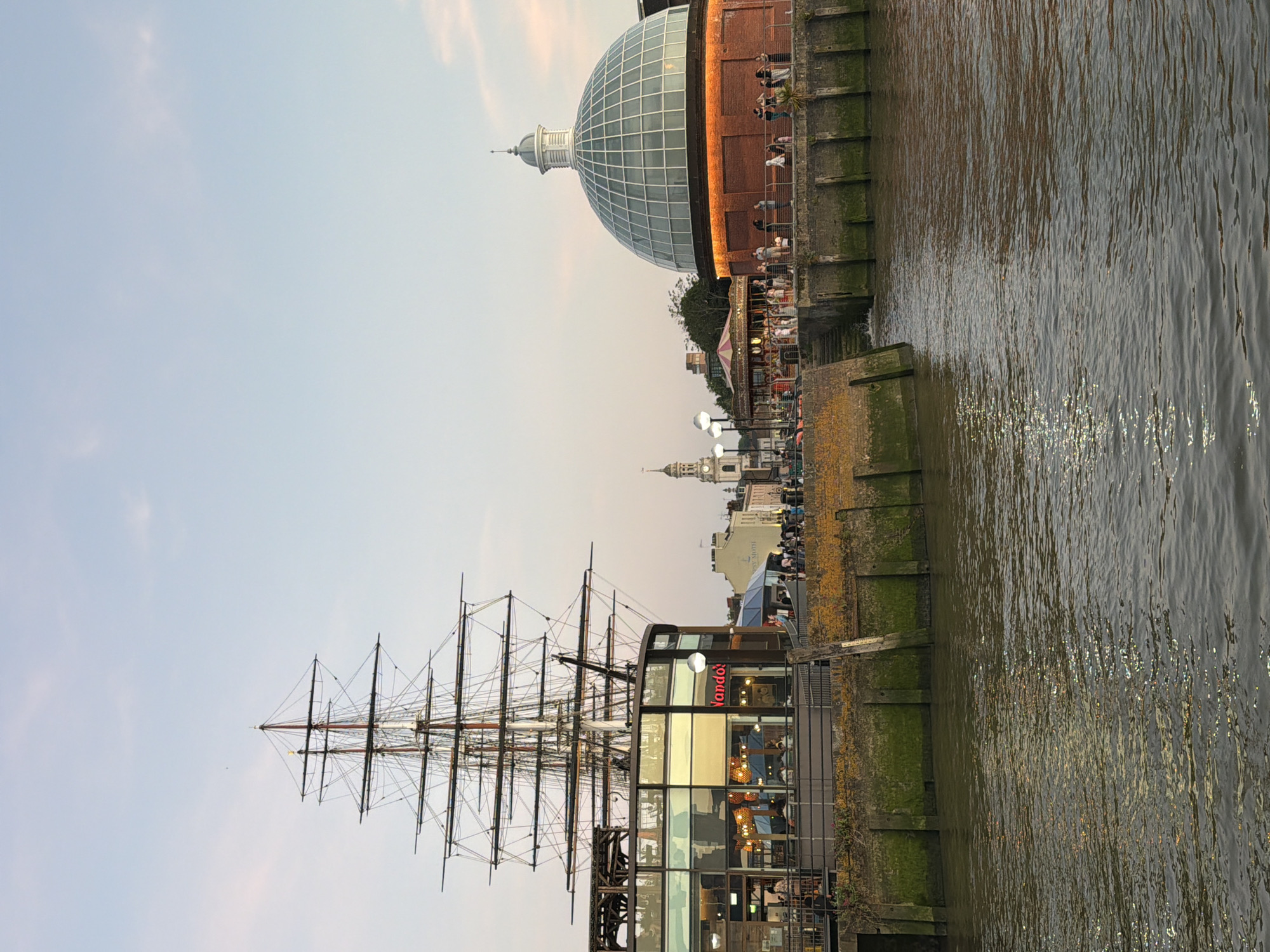 The image features the Cutty Sark, a clipper ship permanently berthed in Greenwich, London, United Kingdom. The scene captures the ship's masts and rigging silhouetted against a pale evening sky. Adjacent to the ship is a modern building with reflective windows, possibly a restaurant. In the background, a large glass-domed building is visible, along with other structures of Greenwich. The foreground shows a section of the wharf or dock with moss or lichen growth, and water with reflections suggesting a river or harbor. There are a few people visible.