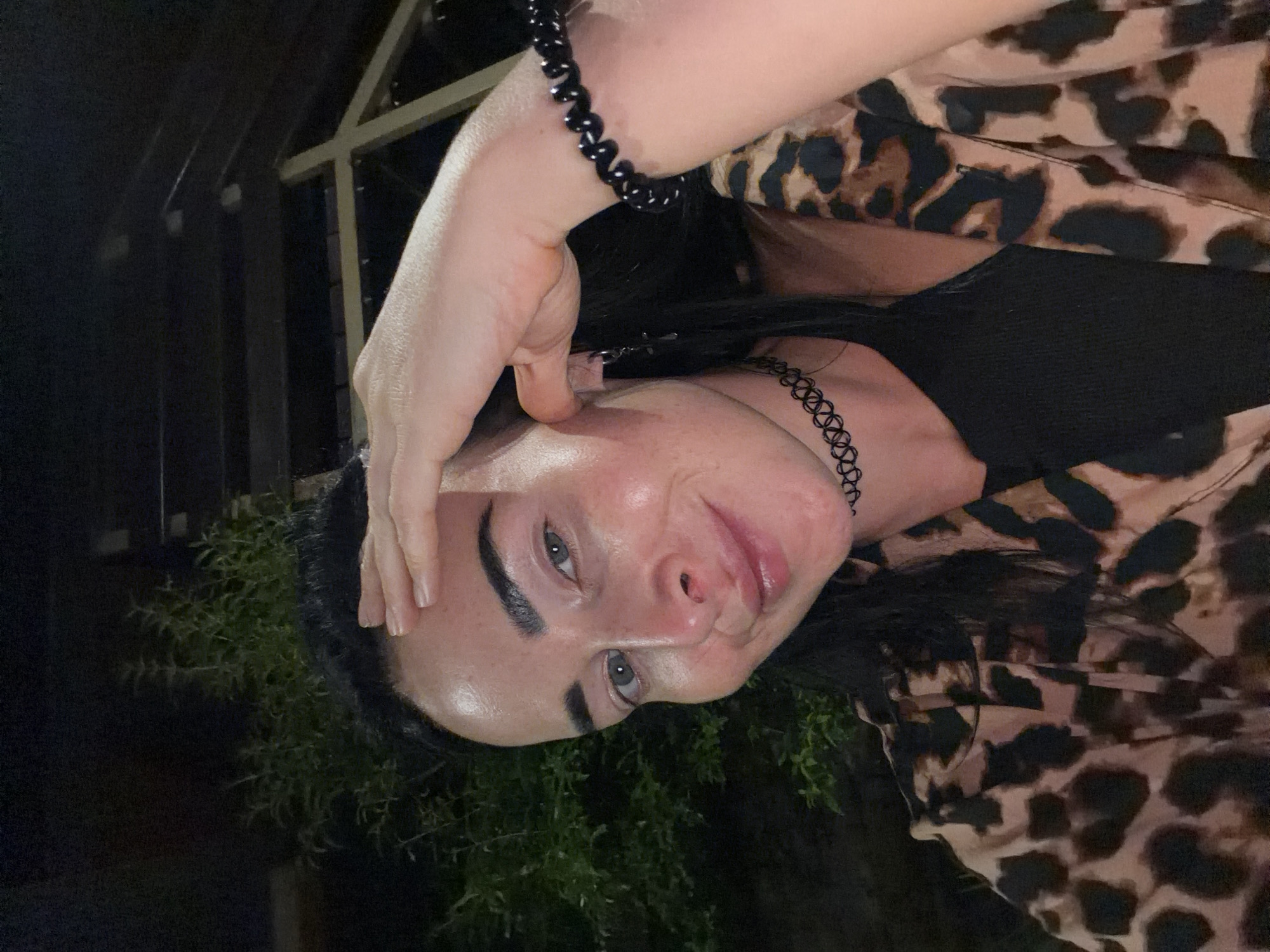 A woman with dark hair and light skin is taking a selfie at night, outdoors. She is wearing a black tank top, a leopard print shirt open over it, a black choker necklace, a black beaded bracelet, and a cross earring. Her right hand is raised to her forehead. The background is dark, with some greenery visible to the left and what appears to be a wooden structure in the upper right quadrant. The image does not contain any identifiable landmarks or clues to suggest a specific location within Sochi, Russia.