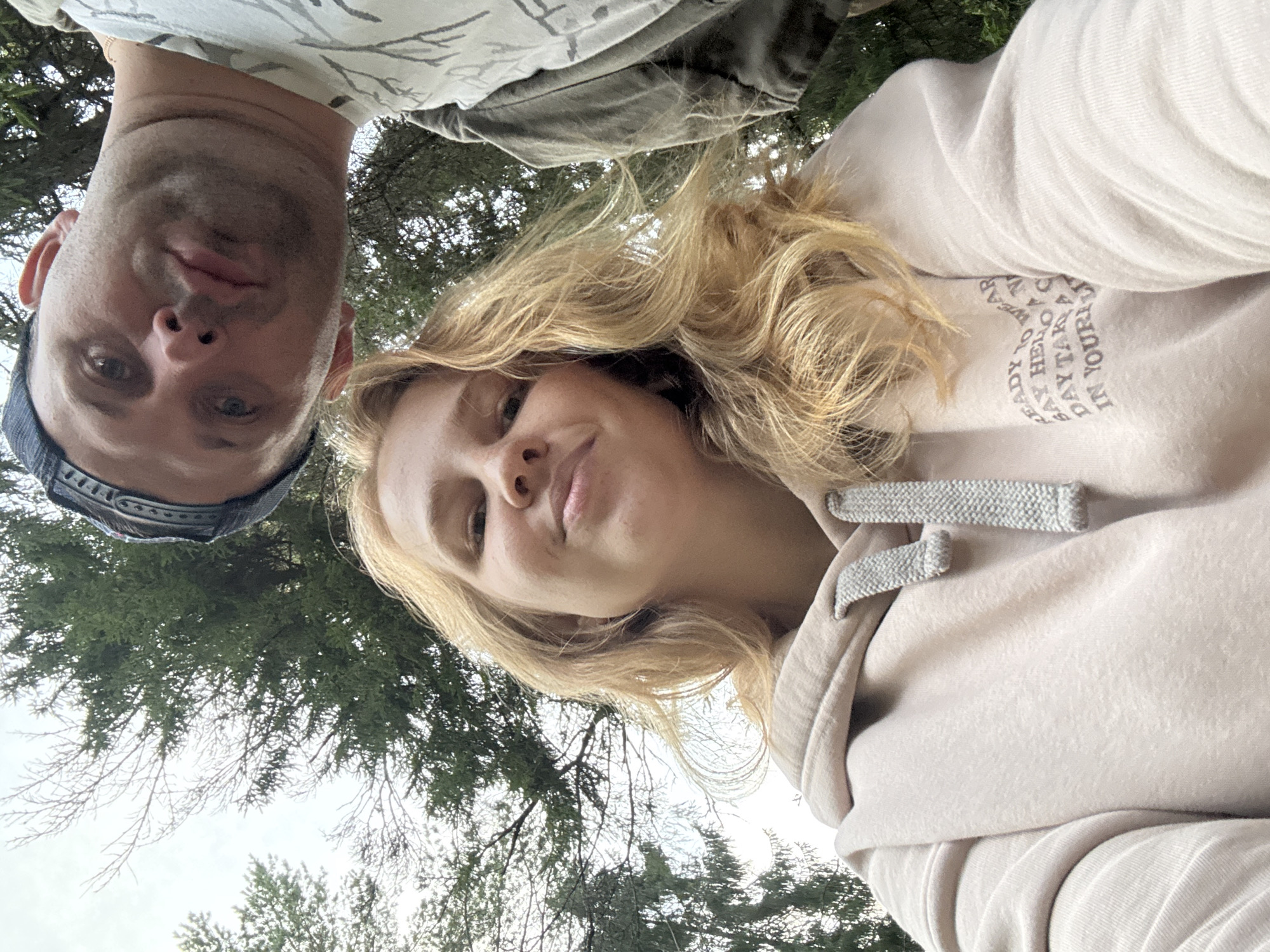 A low-angle, close-up selfie shows two individuals outdoors, with trees and a cloudy sky in the background. The person in the foreground, a young woman with blonde hair, is wearing a light pink hoodie with grey drawstrings. On the front of the hoodie, there is text that reads "READY TO WEAR SAY HELLO DAY TAKE IN YOUR LI". To the right and slightly behind her, a man wearing a blue baseball cap and a t-shirt with a white pattern is visible. Their faces are looking towards the camera. The overall impression is of a casual outdoor setting, potentially a park or wooded area.