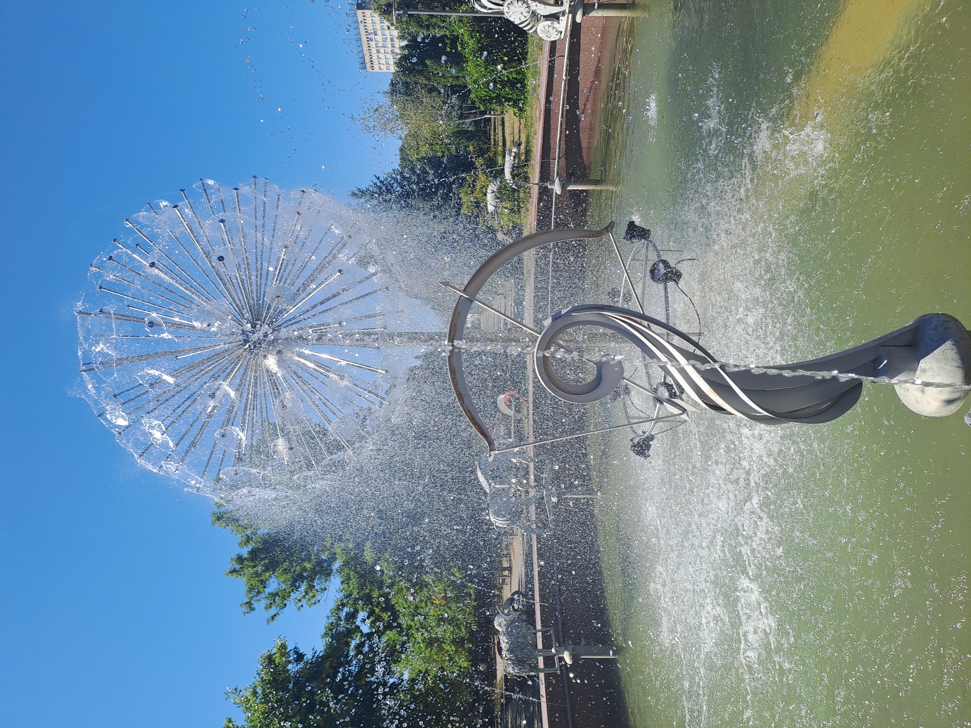 The photograph captures a close-up view of a modern, spherical fountain sculpture spraying water in a park on a sunny day. The sculpture, made of metallic rods, resembles a dandelion seed head or a Sputnik satellite. Water streams outwards from its center, creating a misty effect. In the background, a shallow pool of greenish water reflects the sunlight. Beyond the pool, trees with lush green foliage are visible, and further in the distance, a multi-story building stands under a clear blue sky. Several stylized, gray animal sculptures, possibly sheep or goats, are positioned around the edge of the fountain's basin. The overall scene is one of a public park or recreational area, likely in Chișinău, Moldova, given the specific context provided.