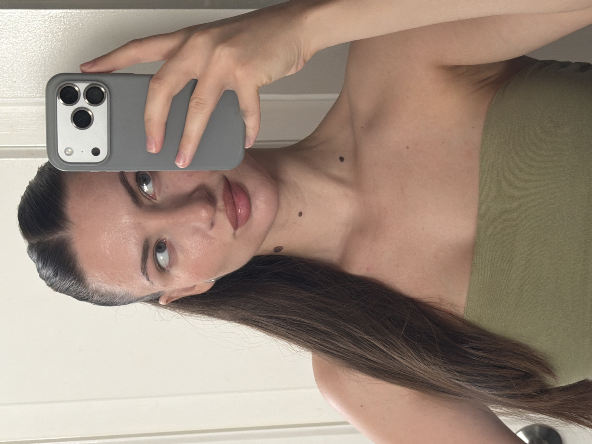 A woman is taking a selfie in what appears to be a bathroom, as indicated by the doorframe visible in the background. She has dark brown hair pulled back into a high ponytail and is wearing a green strapless top. The woman is holding a gray smartphone up to take the picture, which is pointed towards a mirror. Her left eye is looking towards the camera, while her right eye is looking slightly away from the camera. The image does not provide any information that would suggest the location is Sharm El Sheikh, Egypt.