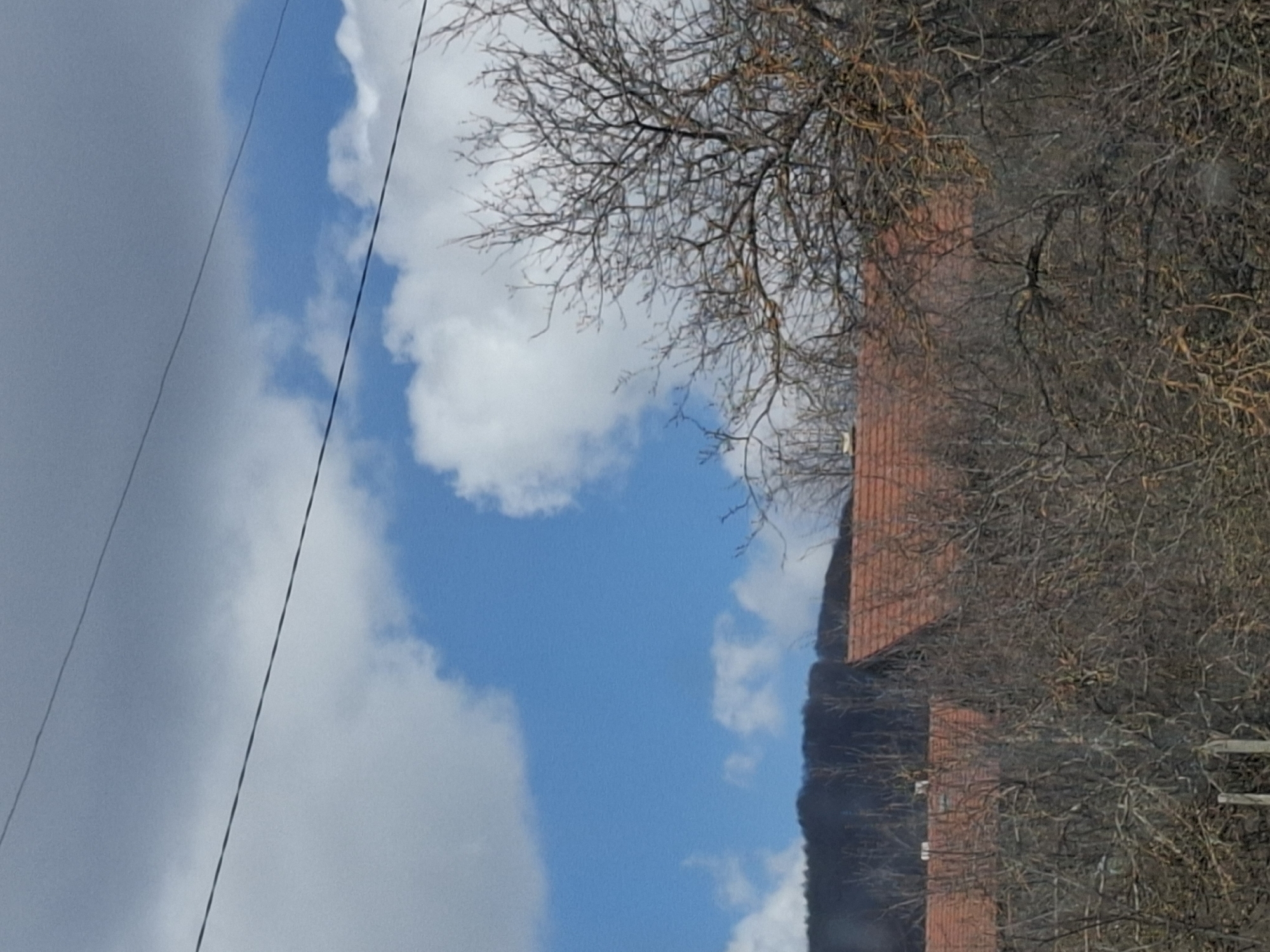 The image shows a view of Gintsi, Bulgaria. The sky is partially cloudy, with a mix of blue sky and gray clouds. A building with a red tile roof is partially visible on the right side of the frame, obscured by bare trees. Utility lines are seen on the left against the sky.