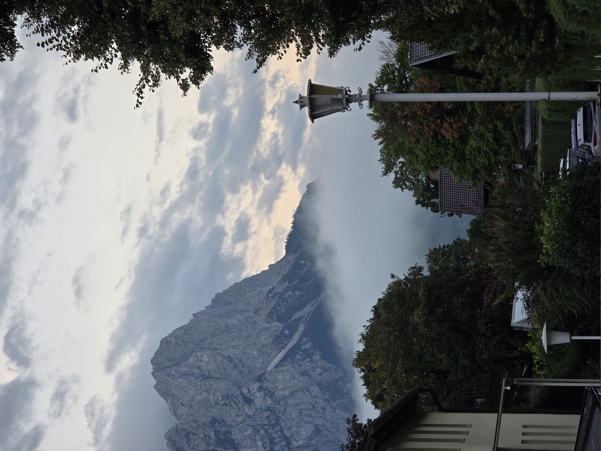 The image shows a view of the Zugspitze mountain range in Garmisch-Partenkirchen, Germany, with low-lying clouds obscuring the base of the mountains. In the foreground, there is a street with a lamppost and several parked cars, including a white caravan and a grey SUV. Houses with tiled roofs are visible amongst lush green trees. The sky is overcast with scattered clouds, and the overall lighting suggests it is either early morning or late afternoon.
