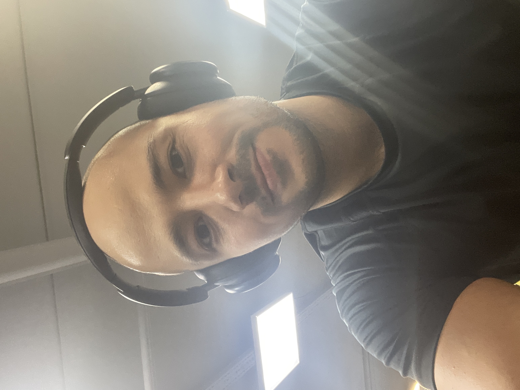 A man with a shaved head and stubble is wearing headphones. He is wearing a gray t-shirt. He is looking down at the camera. There are fluorescent lights on the ceiling.
The location in the city of Izhevsk, Russia cannot be determined from the image.