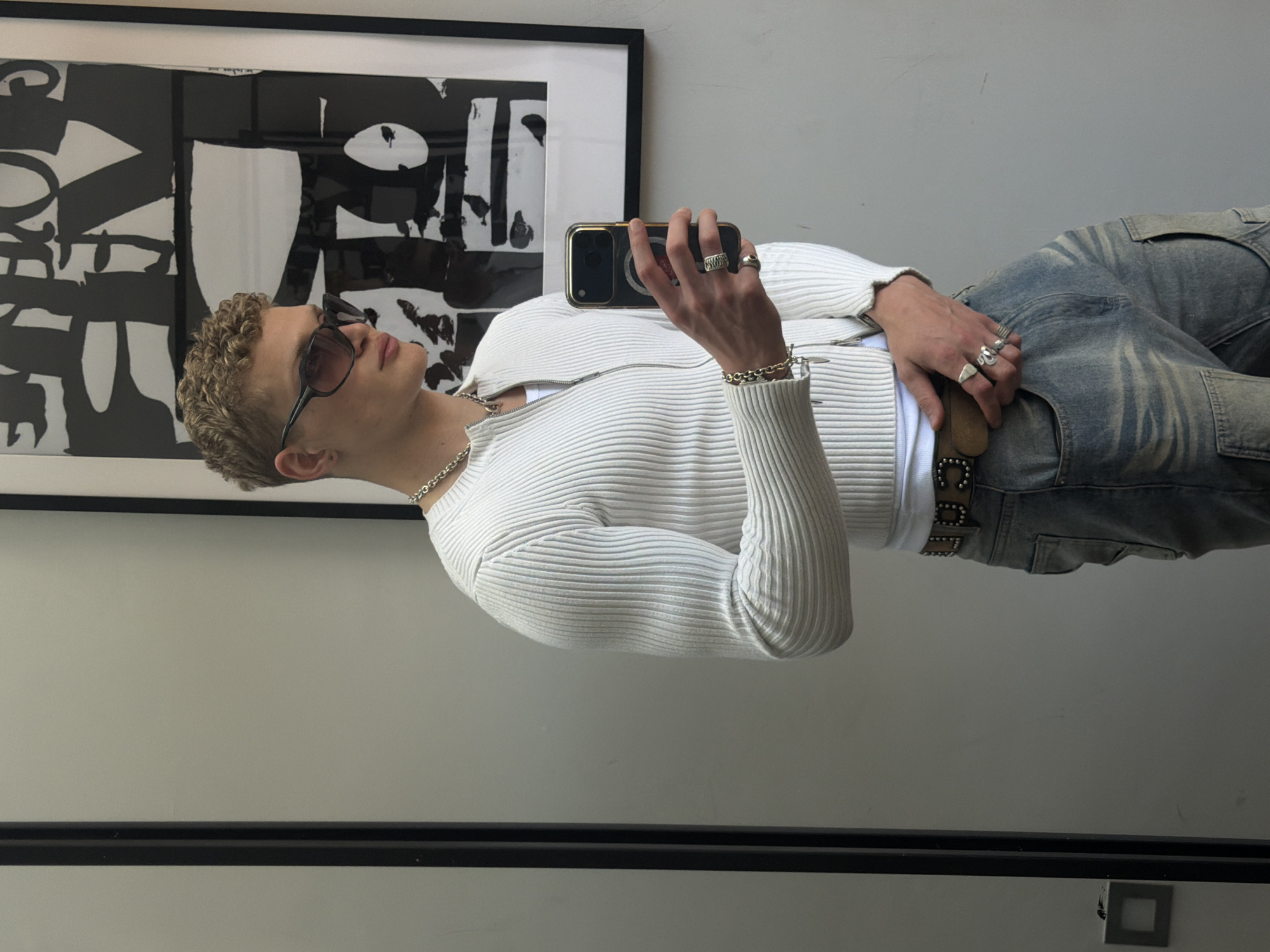 A fair-skinned person with curly blonde hair, sunglasses, a white ribbed cardigan over a white shirt, light blue jeans, and a studded belt is taking a selfie with their phone. The person is wearing jewelry, including a chain necklace, bracelet, and multiple rings. Behind the person is a framed black and white abstract artwork. The scene is indoors with light grey walls. The location appears to be in a modern apartment in London, United Kingdom.