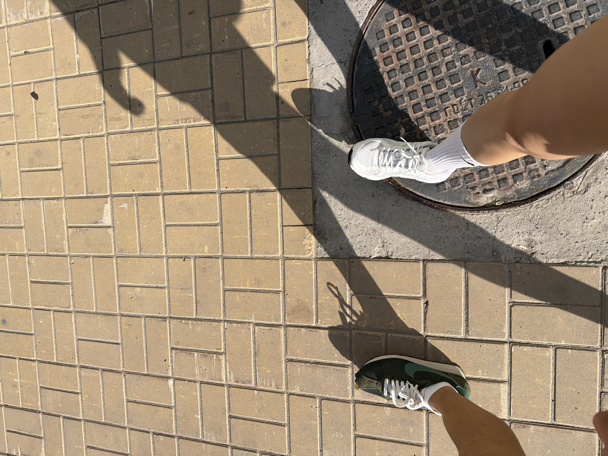 A street-level view shows a person standing on a sidewalk in Tyumen, Russia, with one foot on a manhole cover. The person is wearing different shoes on each foot, one white sneaker and one dark green sneaker. The sidewalk is composed of interlocking tan brick pavers. Shadows of the person are cast across the pavers and the manhole cover.