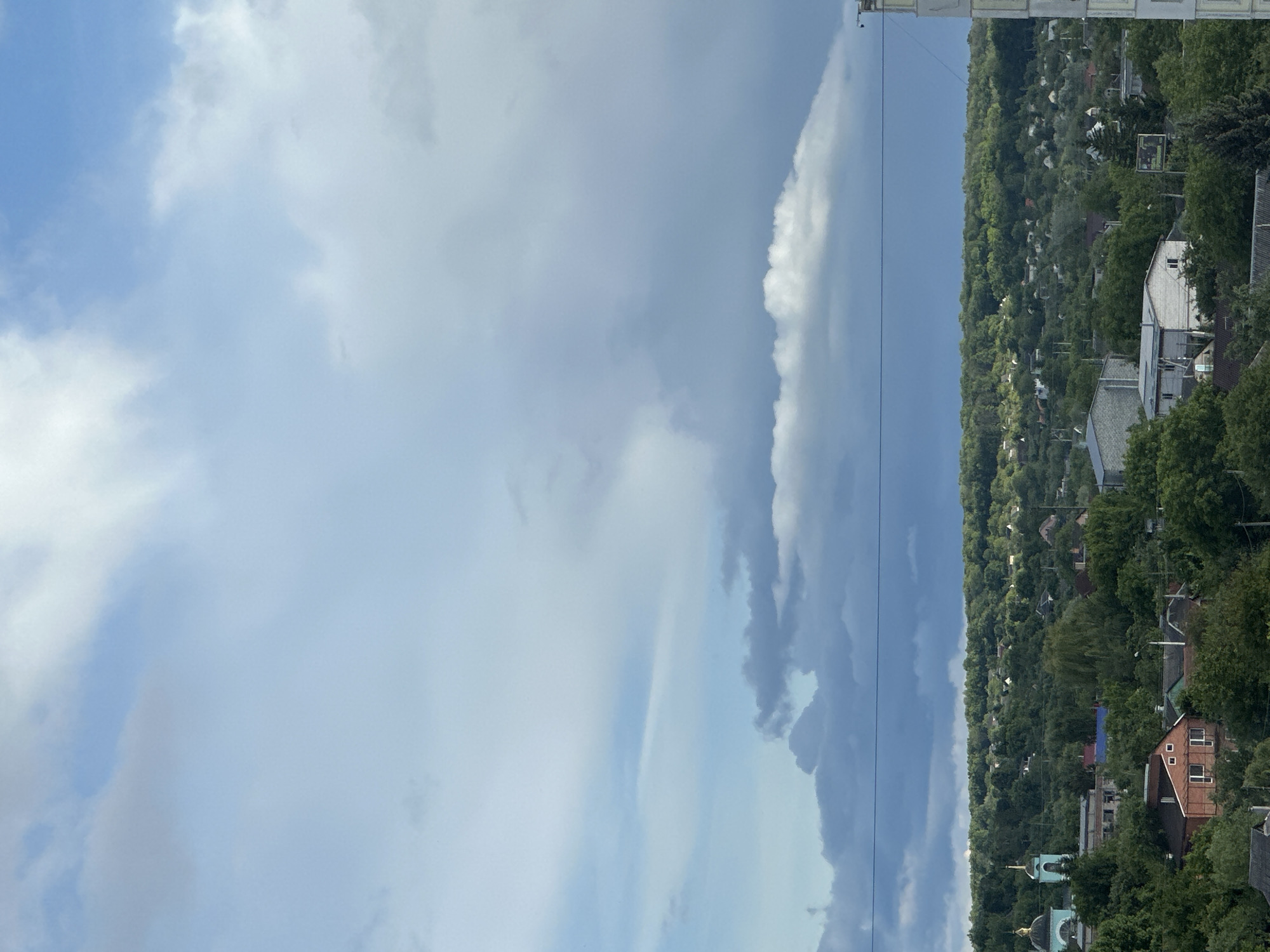 The image shows a view of a cityscape under a cloudy sky. The scene includes houses and trees, with a church visible in the foreground. The clouds range from light blue to dark gray, suggesting potential rain or a storm. The location is Unknown, Unknown.