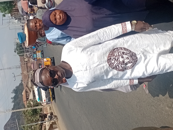 Three people are the focus on a sunny city street. One man wears a white, long-sleeved shirt with a decorative Medusa-like emblem and an ornate pattern on the cuffs, accessorized with sunglasses, a striped cap, and a gold wristwatch. Behind him, a man in a light blue shirt and a woman in a dark blue head covering are visible. The background includes buildings, power lines, a white truck, yellow tricycle taxis, and a mountain in the distance.