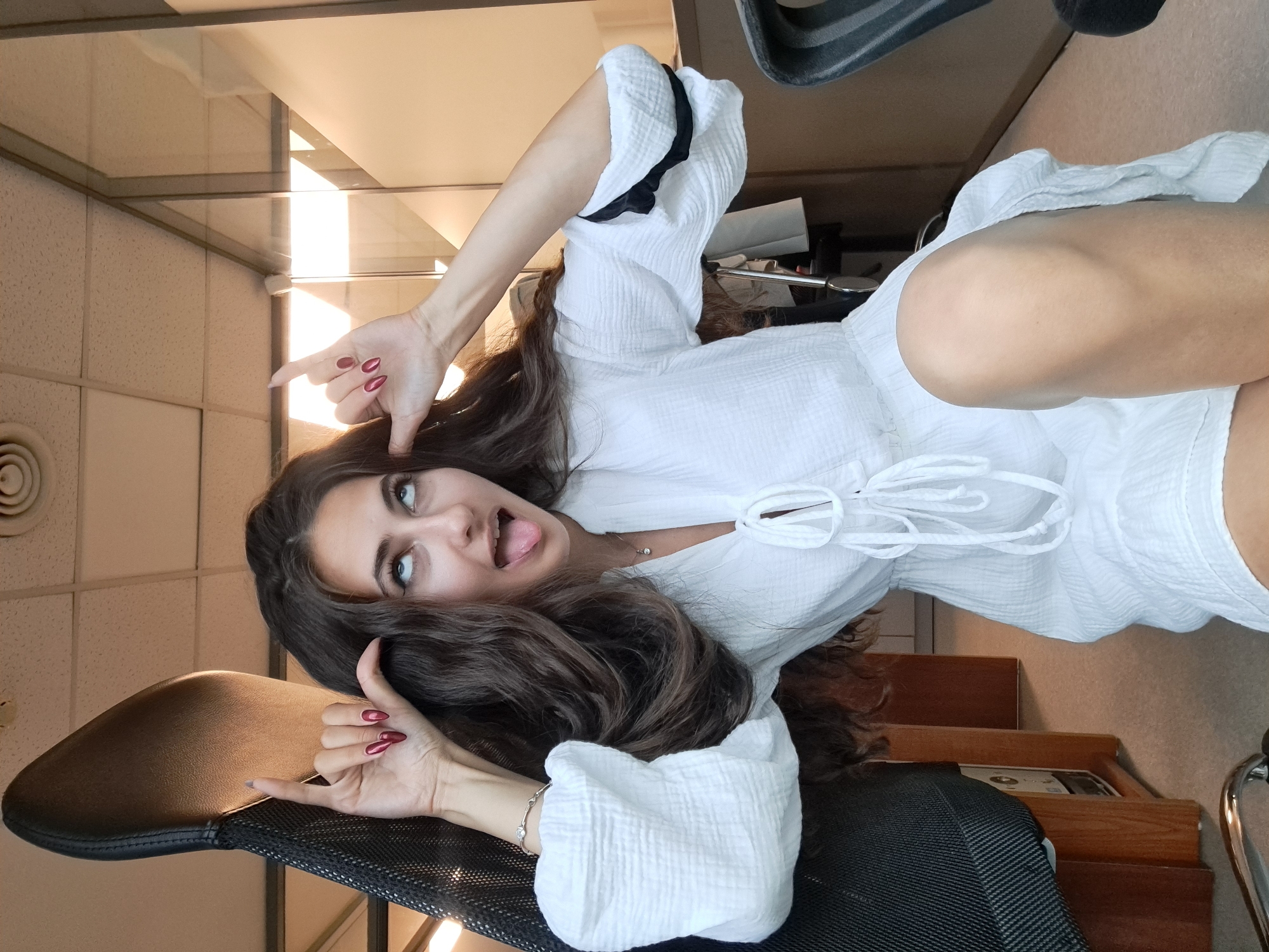 A woman with long, dark hair is sitting in a black office chair. She is wearing a white dress. The woman is sticking out her tongue and making hand gestures resembling devil horns with both hands. The scene appears to be in an office, judging from the ceiling tiles and the desk. The location in the city of Sofia, Bulgaria is not discernible from the image.