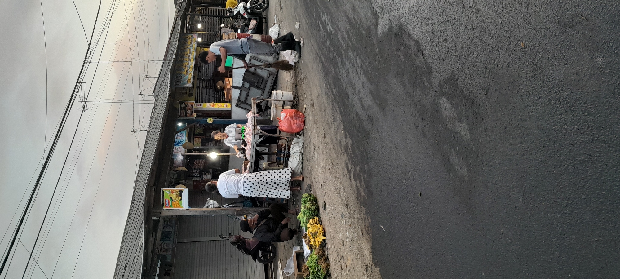 The image captures an outdoor scene at a market in Rogojampi, Indonesia. A market stall with a corrugated metal roof is visible, displaying various goods. In the foreground, a pile of bananas and green vegetables sits on the ground. A man wearing a dark jacket and cap is crouched on the left, observing the scene. In the center, two women are interacting at a table where chickens are being prepared. One woman, wearing a white shirt and a long skirt with black polka dots, stands facing away from the camera. The other woman, in a checkered shirt, is on the right side of the table, holding a small scale and appearing to handle poultry. Behind them, a man in a gray t-shirt and black boots is sweeping the ground with a broom. A large red plastic bag is also on the ground near the table. In the background, the market stall is filled with various items, including what appears to be food products. Motorcycles are parked to the left and right of the stall. The sky above is overcast with power lines crisscrossing the frame. The ground is a dark, wet asphalt surface.