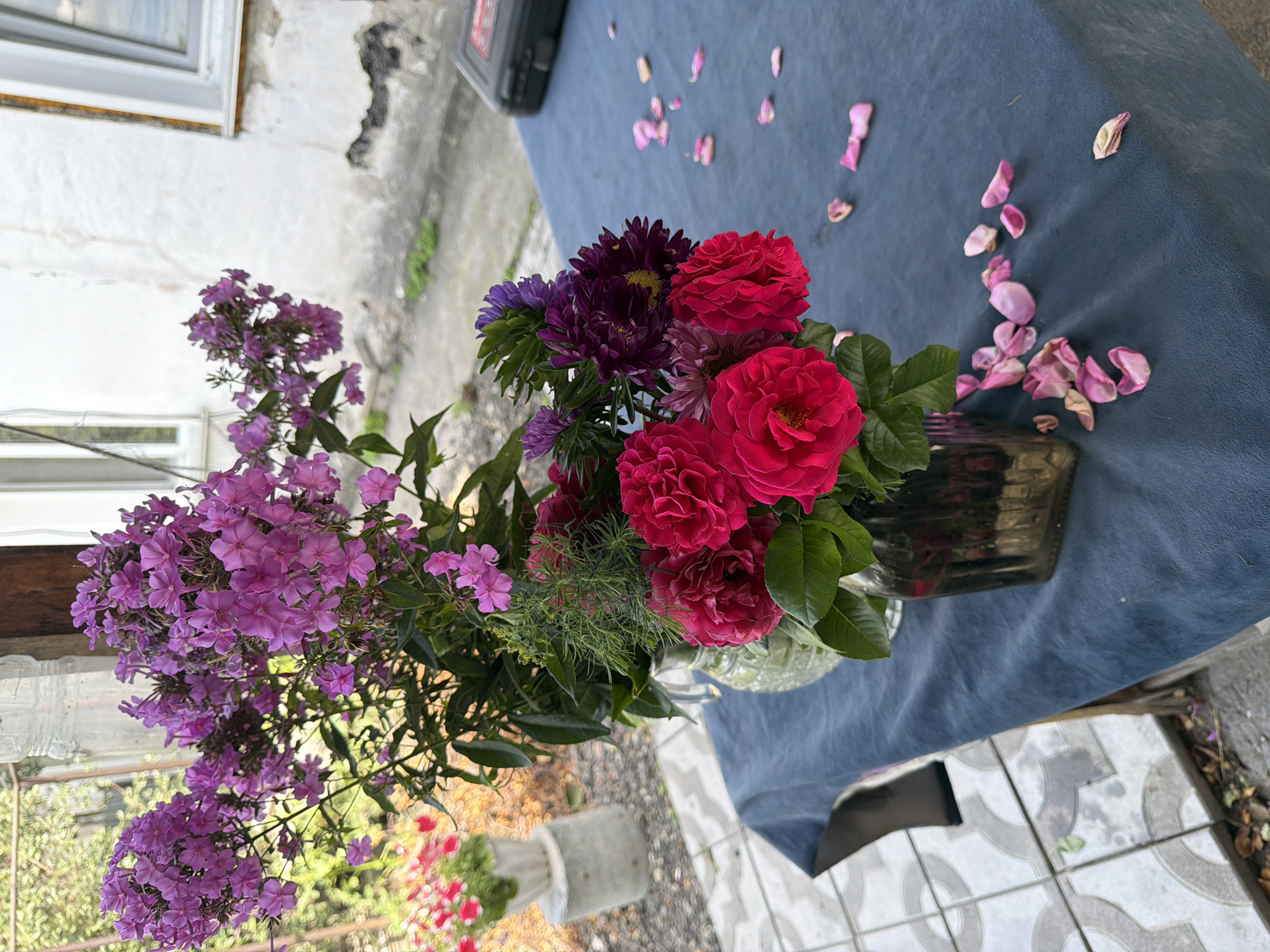 A still life photograph captures a table covered with a dark blue tablecloth, set outdoors in what appears to be a yard or courtyard. On the table, two glass vases hold a vibrant bouquet of flowers. The larger vase on the left contains tall, cascading arrangements of pink and purple phlox, interspersed with delicate green foliage. To the right, a more compact vase holds a cluster of bright red roses and deep purple asters. Scattered across the tablecloth are several fallen rose petals, adding a touch of ephemeral beauty to the scene.
In the background, a white building with visible peeling paint and cracked plaster suggests an aged structure. A window with white trim is visible on the building's facade. To the left of the table, a concrete pedestal supports a pot with pink flowers, adding another splash of color to the outdoor setting. The ground visible beyond the table is paved with light-colored tiles, some of which appear to be stained or weathered. The overall impression is one of a tranquil, possibly rustic, outdoor domestic space. The presence of a dark rectangular object, possibly a toolbox, on the far right of the table hints at practical activities associated with the location. The lighting suggests it is daytime, with diffused sunlight illuminating the scene. The location is specified as Semey, Kazakhstan.