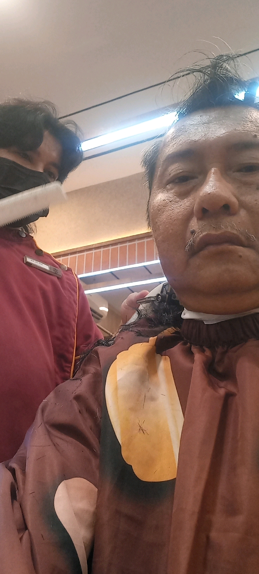 In City of Medan, Indonesia, two individuals are present in what appears to be a barbershop. The individual on the right is a man with dark hair, a mustache, and is wearing a brown cape with a yellow abstract design. He is in the process of having his hair cut. The individual on the left is wearing a dark face mask and a maroon uniform with a name tag. They are holding a comb. The background shows a ceiling with fluorescent lighting and a wall with vertical tiles. The lighting is bright, suggesting an indoor setting.