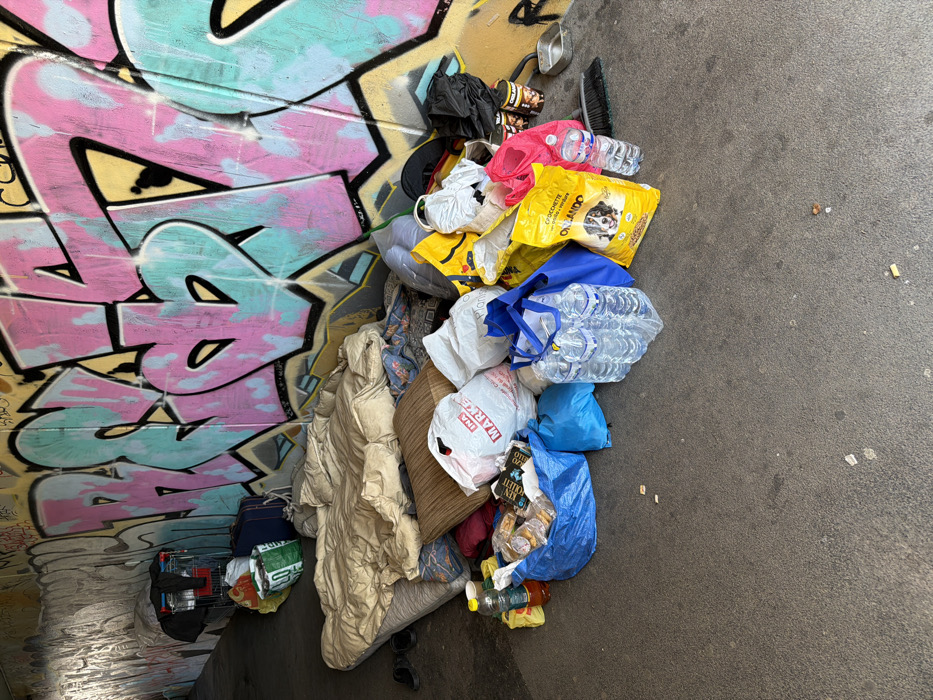 The scene depicts a makeshift encampment within an urban underpass or tunnel in Milan, Italy. The concrete wall on the right is extensively covered in vibrant graffiti, primarily featuring large letters and shapes in pink, light blue, yellow, and black outlines, including elements resembling hearts. The floor is a grey concrete surface.
On the left side, further into the tunnel, a metal shopping trolley holds various items, including dark fabric and a red-orange bag. Adjacent to the trolley are two blue suitcases and a green shopping bag.
Central to the foreground, a mattress is laid on the concrete floor, covered with a beige duvet or blanket and a pillow. Two dark flip-flops rest on the ground near the mattress.
To the right of the sleeping area, a collection of provisions and personal items is assembled. This includes numerous plastic and fabric shopping bags, some appearing full; notable brands visible are a white "INA MARKET" bag and a yellow bag of "ORLANDO CROCCHETTE" dog food featuring a dog's image. A six-pack of bottled water is contained in clear plastic, alongside several loose water bottles. Other items include an amber-colored liquid bottle, several cans of food (one labeled "DELANO"), a small metal bowl, and a small broom.
No individuals are present in the image, and no actions or interactions are depicted. The area is illuminated, suggesting either natural light from an opening or artificial lighting.
