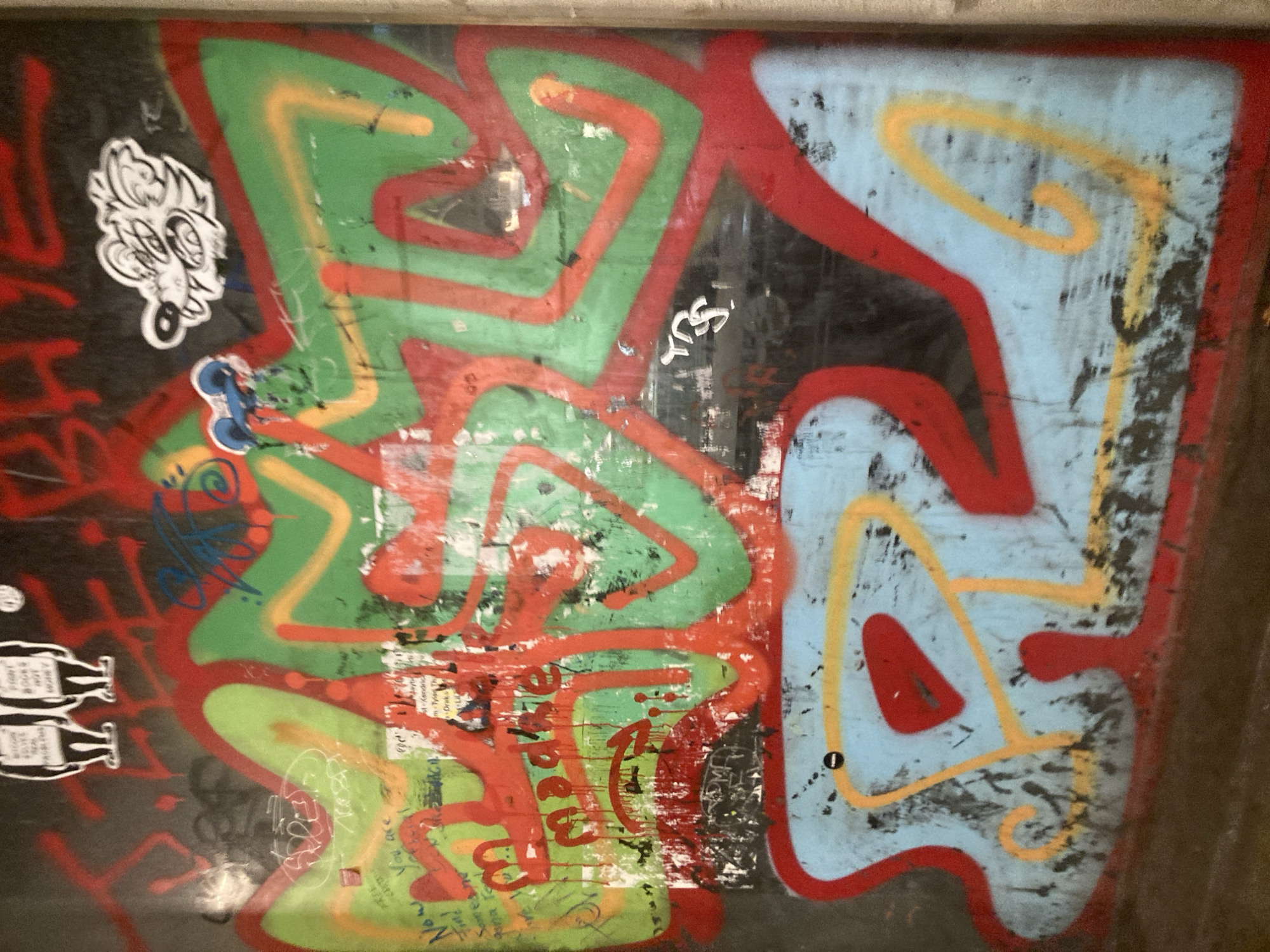 This image captures a section of a wall covered in vibrant graffiti. The graffiti features a mix of spray-painted designs and stickers. Prominent colors include bright red, green, yellow, and light blue. There are stylized cartoonish figures, possibly a wolf-like creature and a two-headed figure wearing hats, along with abstract shapes and lettering. The wall itself appears to be aged and weathered, with visible scuffs, scratches, and some peeling paint or posters underneath the graffiti. The lower portion of the image shows a large, stylized blue shape outlined in red, with yellow and red accents within. The setting appears to be an urban environment, possibly an alleyway or a designated graffiti wall, though a specific location in Barcelona is not identifiable from the visual information alone. There are no people or active interactions depicted in the image.