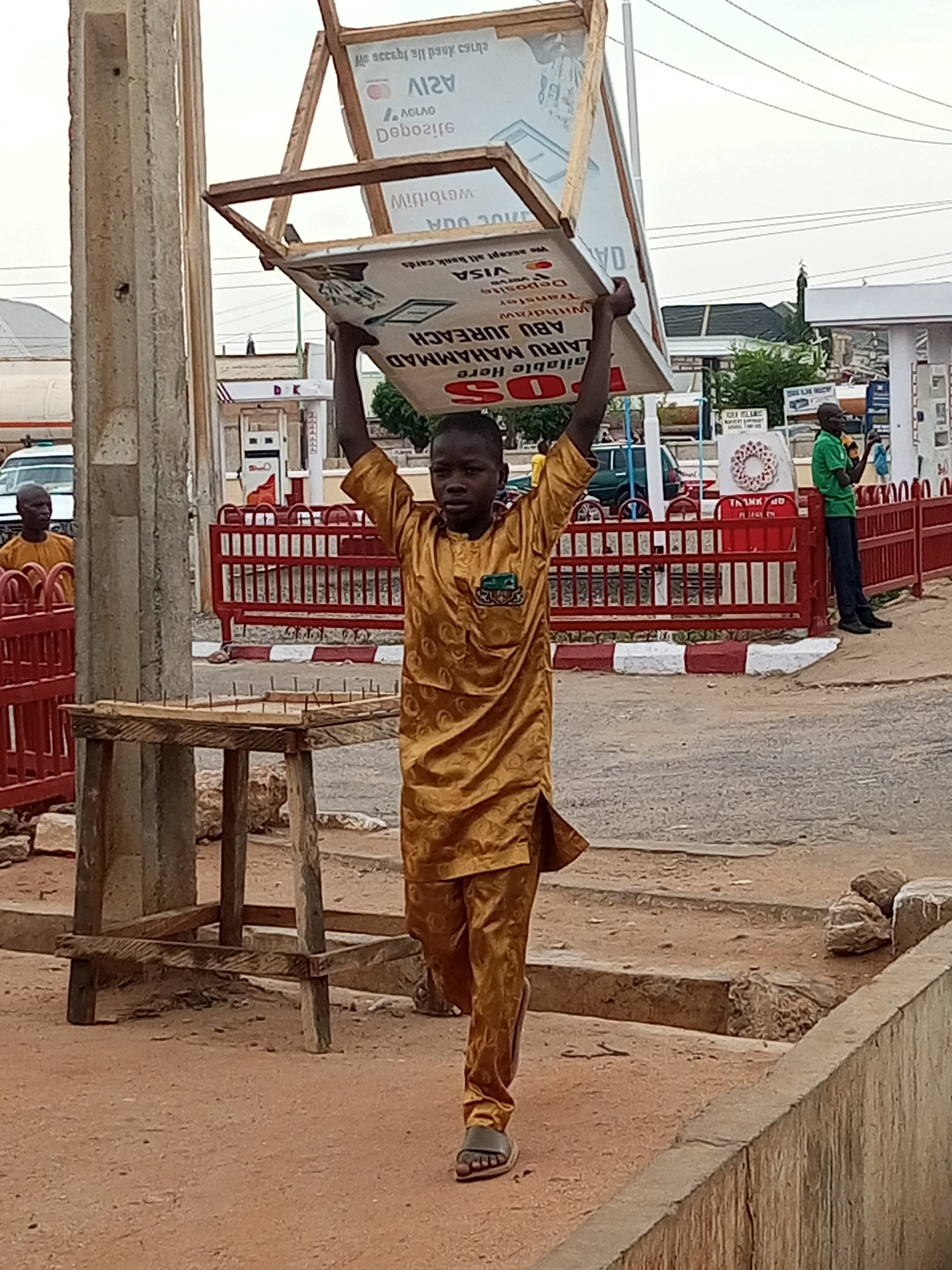 A young person walks on a dirt and concrete surface, wearing a brown, long-sleeved shirt and pants, and sandals. On their head, they balance a large sign with text in English and other languages, supported by a wooden frame. A wooden table with nails sticking up from the surface sits to the left of the person. Red metal railings line the street. In the background, a gas station is visible with a man in a green shirt standing near the red railings, and another person wearing a brown shirt standing by the red railings. The image was taken during the day in Tumfure, Nigeria.