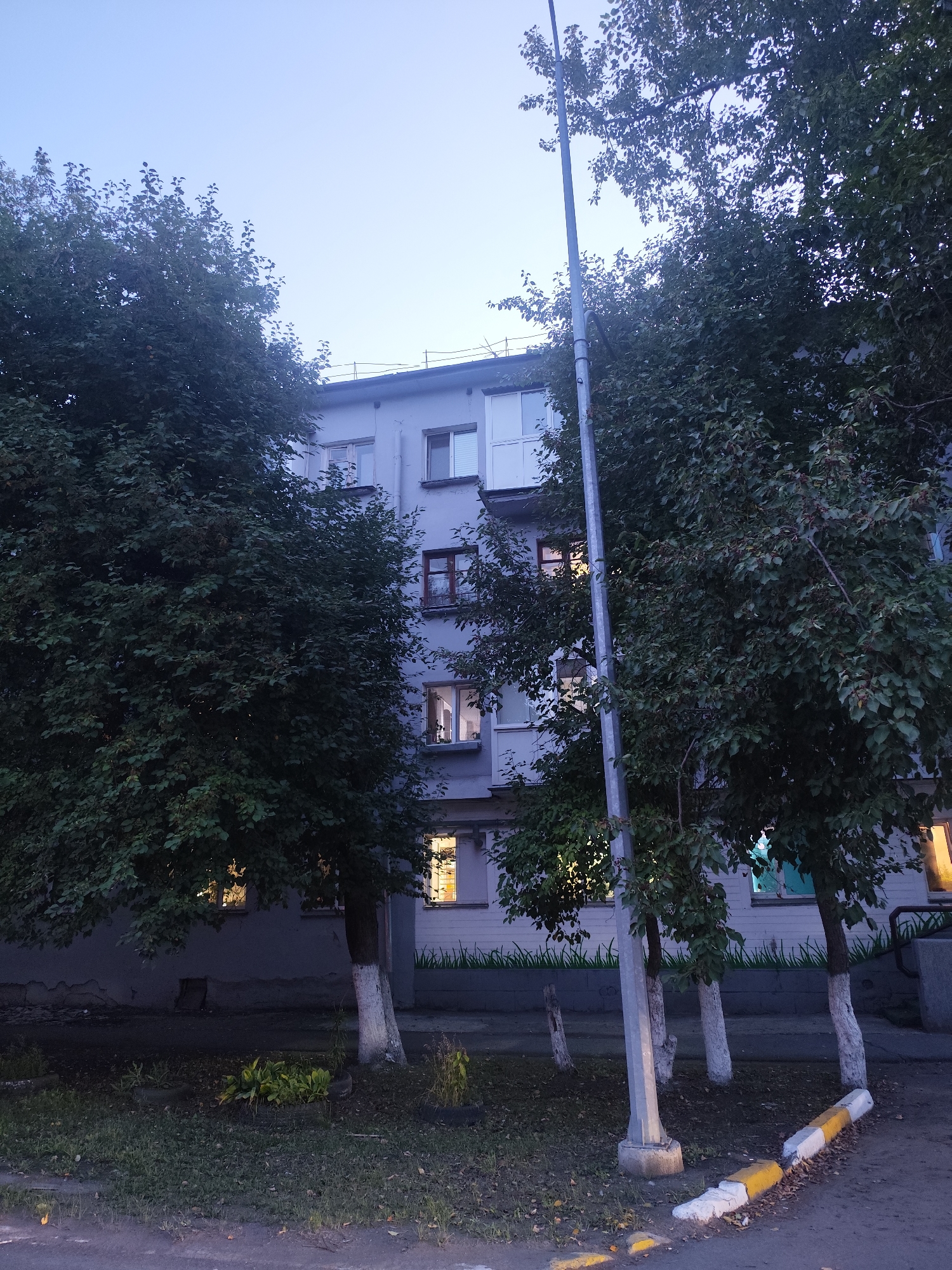 The image depicts a view of a multi-story apartment building in Petropavl, Kazakhstan, during twilight hours. The building appears to be constructed of light-colored materials, possibly concrete or stucco, with several windows visible. Some windows are illuminated from within, casting a warm glow.
In front of the building, several trees with dense green foliage are present, some of which are partially obscuring the building's facade. The trees have their lower trunks painted white, a common practice in some regions for tree preservation or aesthetic purposes. A tall, slender light pole stands vertically in the foreground, positioned between the trees and the building.
The ground in front of the building is covered with grass and some fallen leaves, suggesting an outdoor area like a courtyard or a residential street. There is a paved surface in the very bottom foreground, possibly a sidewalk or road, with yellow and white painted curbs or road markers. The overall atmosphere is subdued due to the low light conditions of dusk. No people or vehicles are visible in the scene.