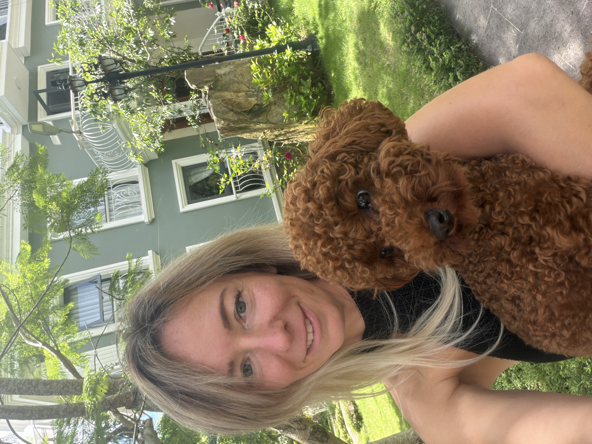 A woman with blonde hair and a black top is holding a small brown poodle. They are outdoors in front of a light green building with white trim. The building has multiple windows and a balcony with a white railing. Trees and green grass are visible in the background. The overall setting appears to be a residential area in Xuân Trường - Đà Lạt, Vietnam, likely a garden or front yard, with ample sunlight.