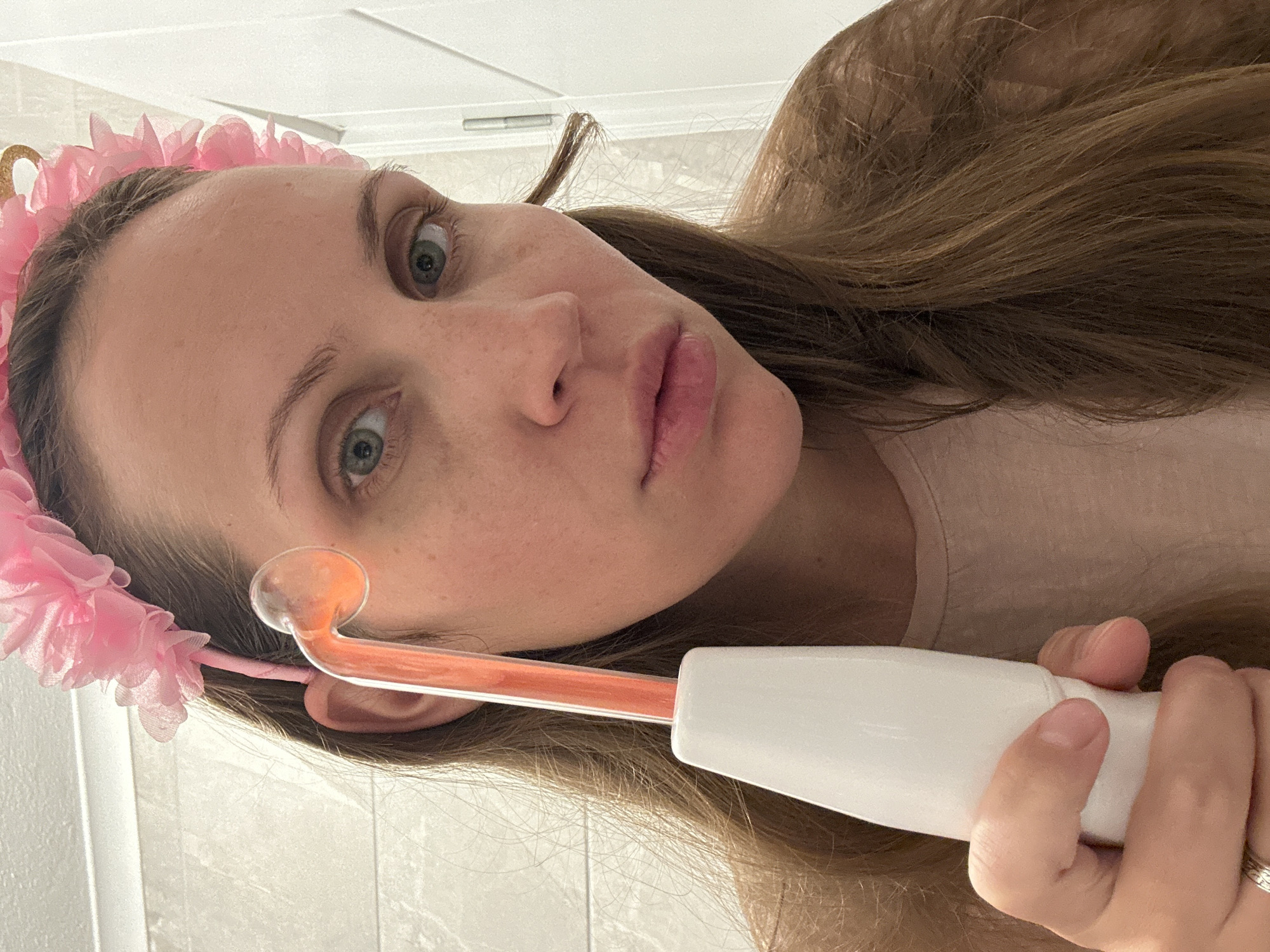 A woman holds a high-frequency facial wand. She is wearing a pink floral headband. She has light skin, light eyes, and light brown hair. The background is blurred and shows a white wall. There is no indication of Fethiye, Turkey in the image.