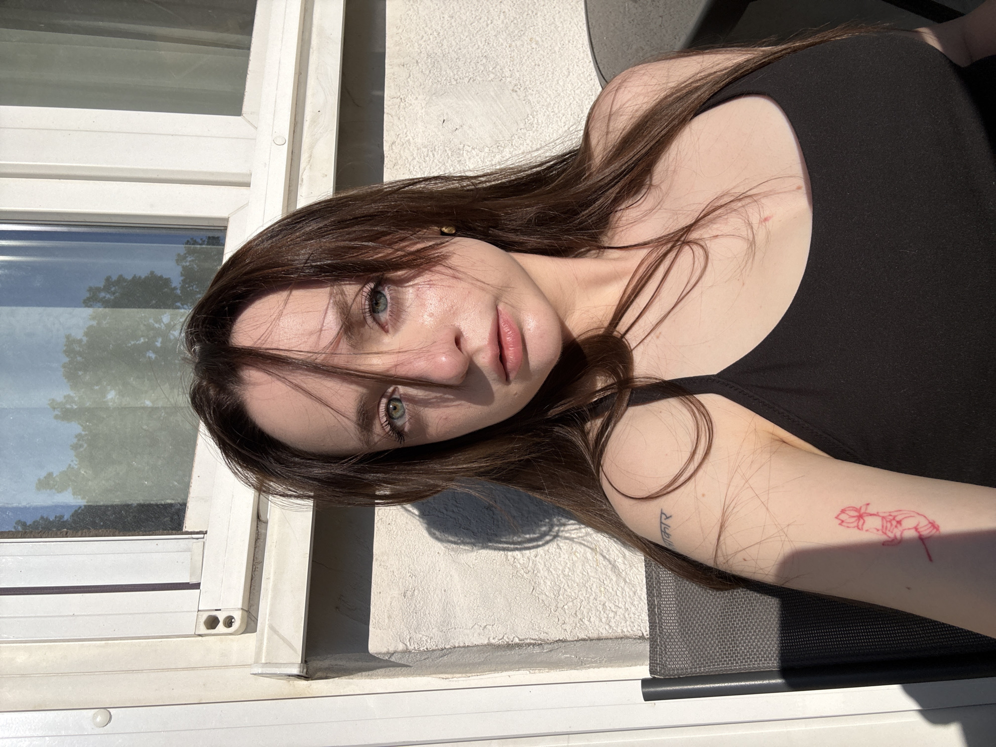 A young woman with long, dark brown hair and green eyes is pictured from the chest up, sitting outside in what appears to be Belgrade, Serbia. She is wearing a black tank top and has a red tattoo of a lotus flower on her left arm, as well as a script tattoo on her left shoulder. The sun is shining brightly, casting a shadow of her on the textured, white wall behind her. A portion of a white-framed window with reflections of trees is visible in the upper background. She is sitting in a dark-colored chair next to a table, the edge of which is visible on the right side of the frame. The lighting suggests it is either early morning or late afternoon.
