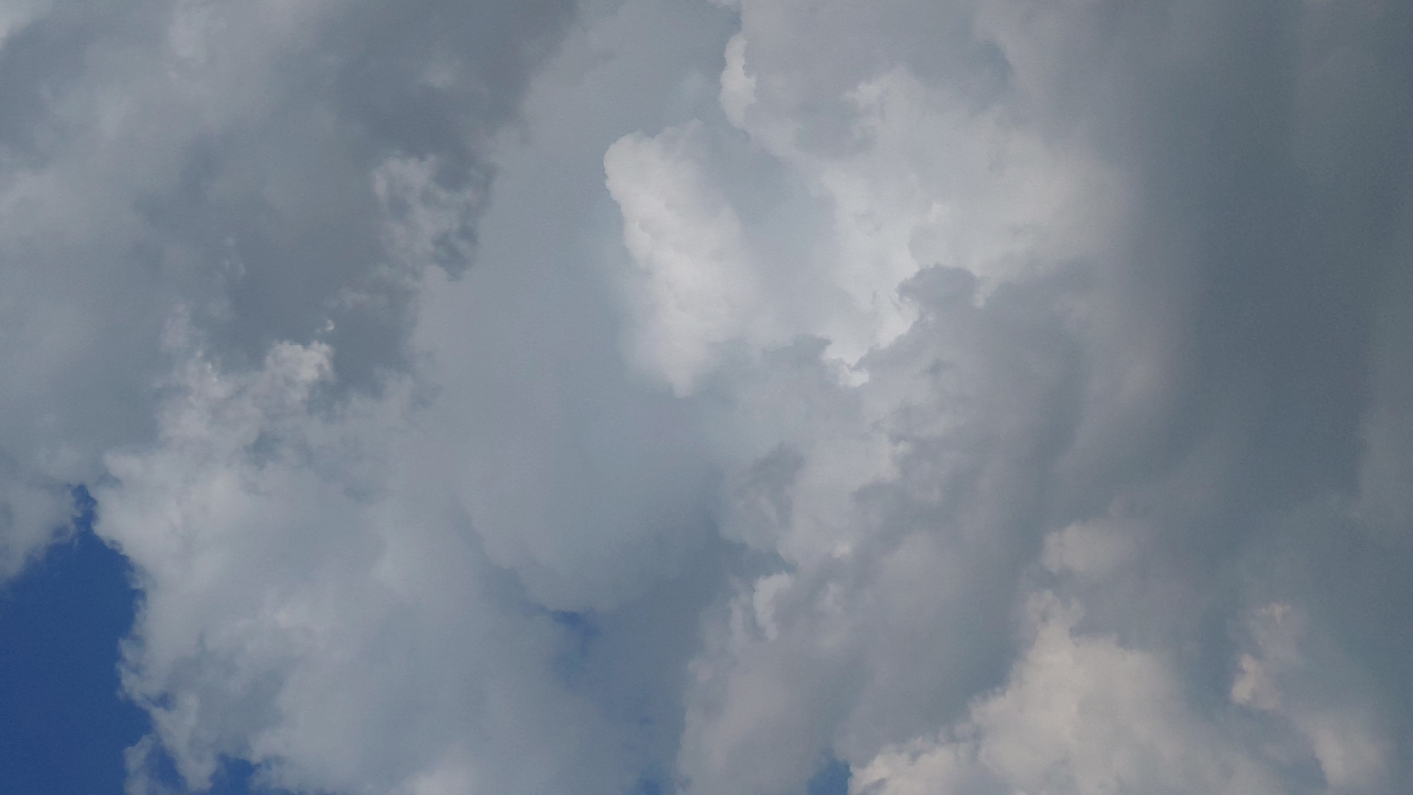The image is a close-up, vertical shot of a cloudy sky. There are patches of bright blue visible in the upper left corner, suggesting it is daytime. The clouds are varied in texture and density, with some appearing wispy and white, while others are darker and more ominous, indicating potential for rain or a storm. The lighting suggests the sun is present but obscured by the clouds, creating a diffused and somewhat dramatic atmosphere. No people, animals, or man-made structures are visible in the frame. The scene depicts a natural sky formation, with no specific indicators of its location within the city of Abovyan, Armenia.