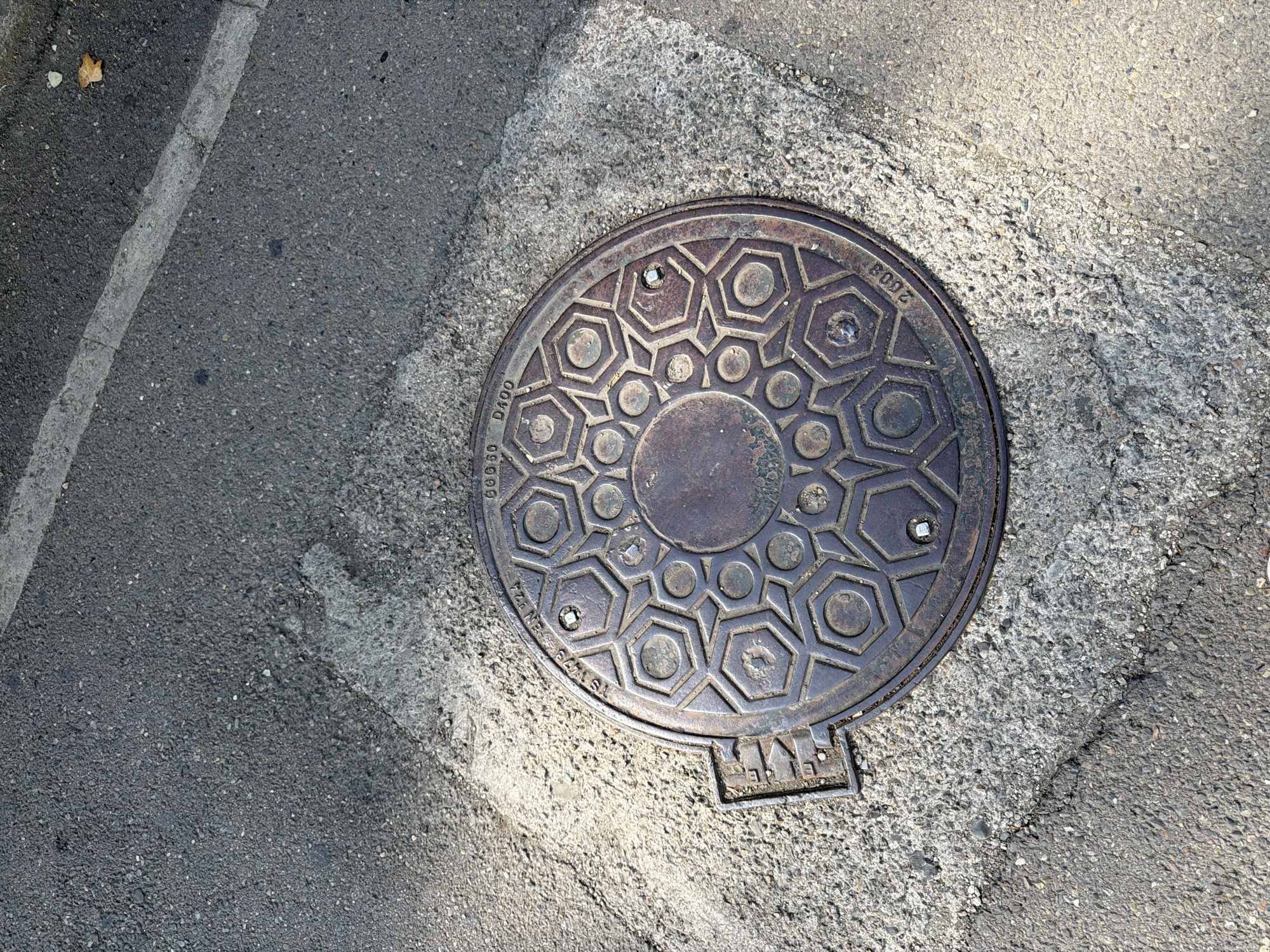 An overhead shot captures a circular, dark metal manhole cover with an intricate hexagonal pattern, set into a concrete and asphalt surface. The manhole cover displays raised lettering that reads "TS 1978 EN 124" and "600x50 D400," along with the year "2008." A small, rectangular metal lever is attached to the left side of the manhole cover. The surrounding ground is a mix of dark asphalt with visible aggregate and lighter-colored concrete, showing cracks and wear. A thin, light-colored line, possibly a road marking, runs diagonally across the upper right portion of the frame. The lighting suggests a sunny day, with shadows cast on the ground. This scene is located on a street in Tbilisi, Georgia, indicated by the distinctive manhole cover markings which align with Georgian standards for manhole covers.