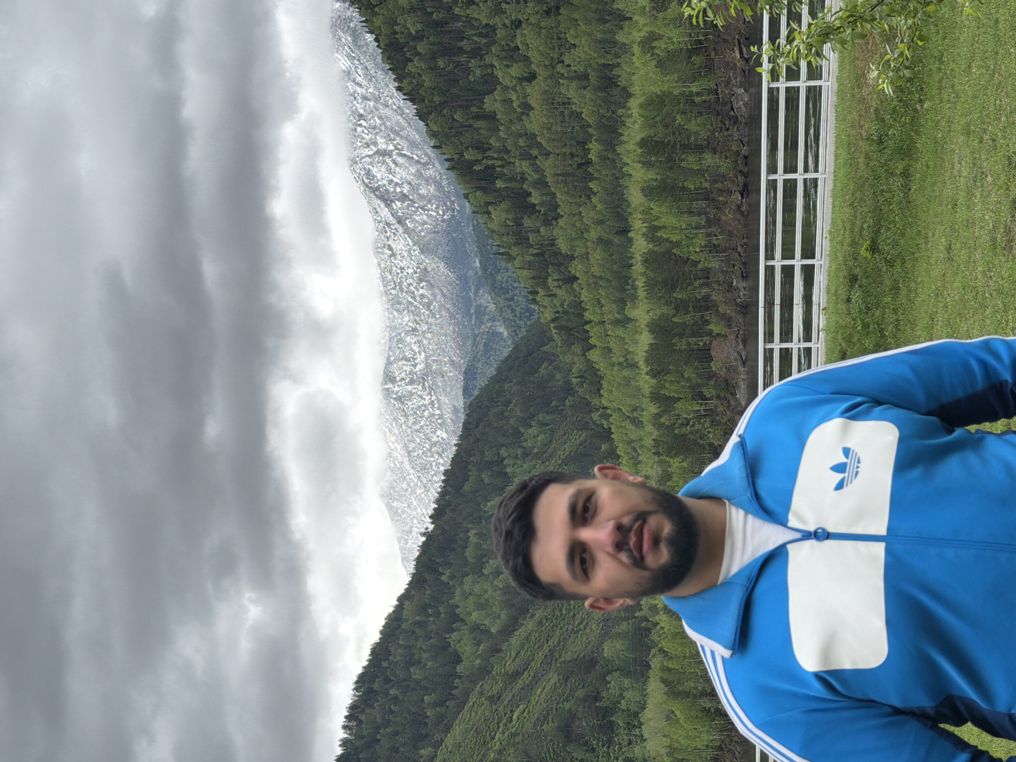 The image shows a man in a blue Adidas tracksuit posing in front of a mountain range. The mountains are partially covered in snow and surrounded by a dense forest. A white fence is visible in the background on the right side of the frame. The sky is overcast with heavy clouds. The location within Cheremushki, Russia, is not identifiable from the image.