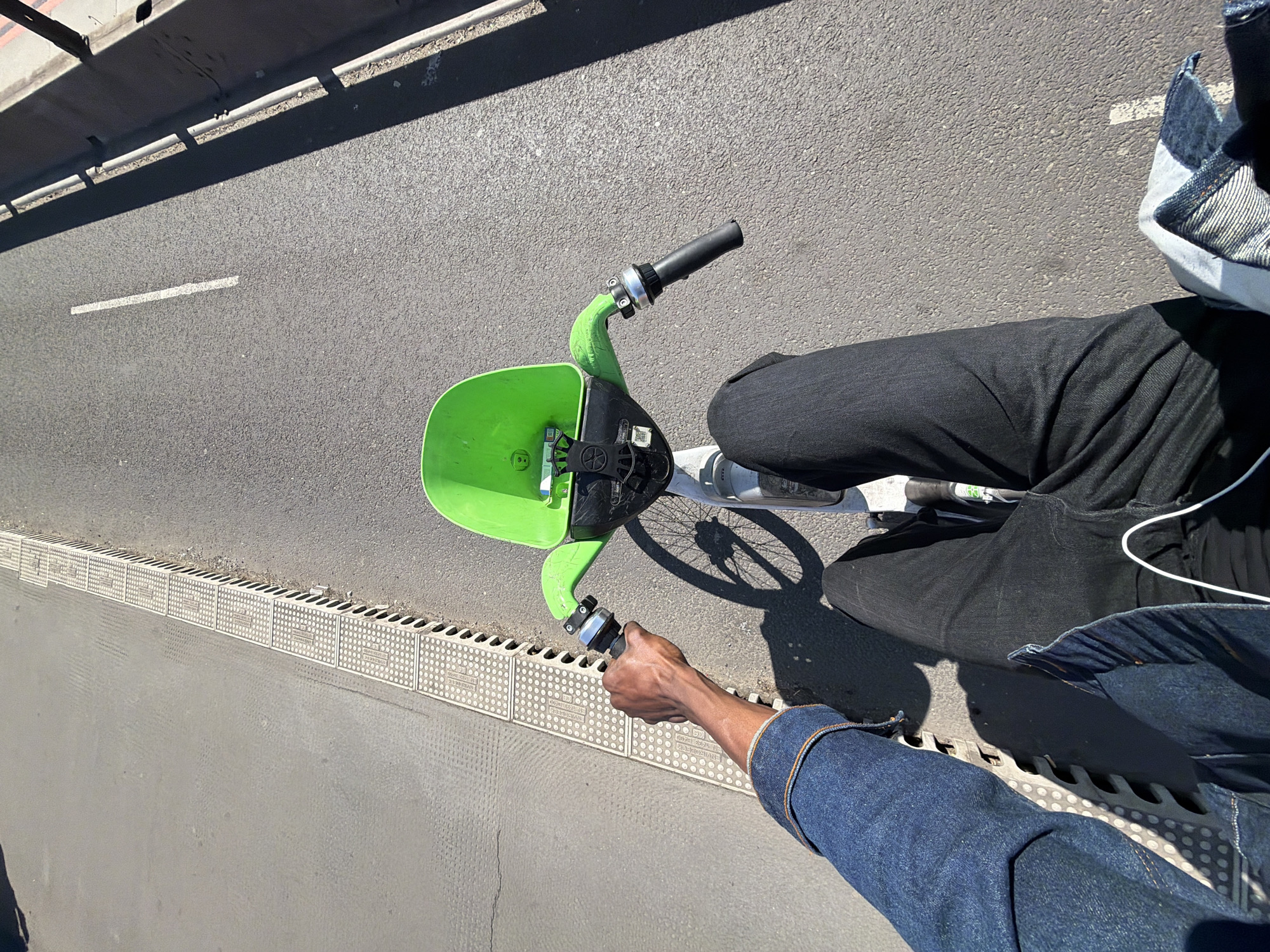 A person is standing next to a lime green Lime e-bike on a street in London, United Kingdom. The person is wearing black pants, a denim jacket, and white sneakers. The person's arm is reaching out to steady the e-bike. The street is gray asphalt with white lines. There are textured stone drainage covers along the edge of the road. There is a shadow cast over the upper left corner of the image.