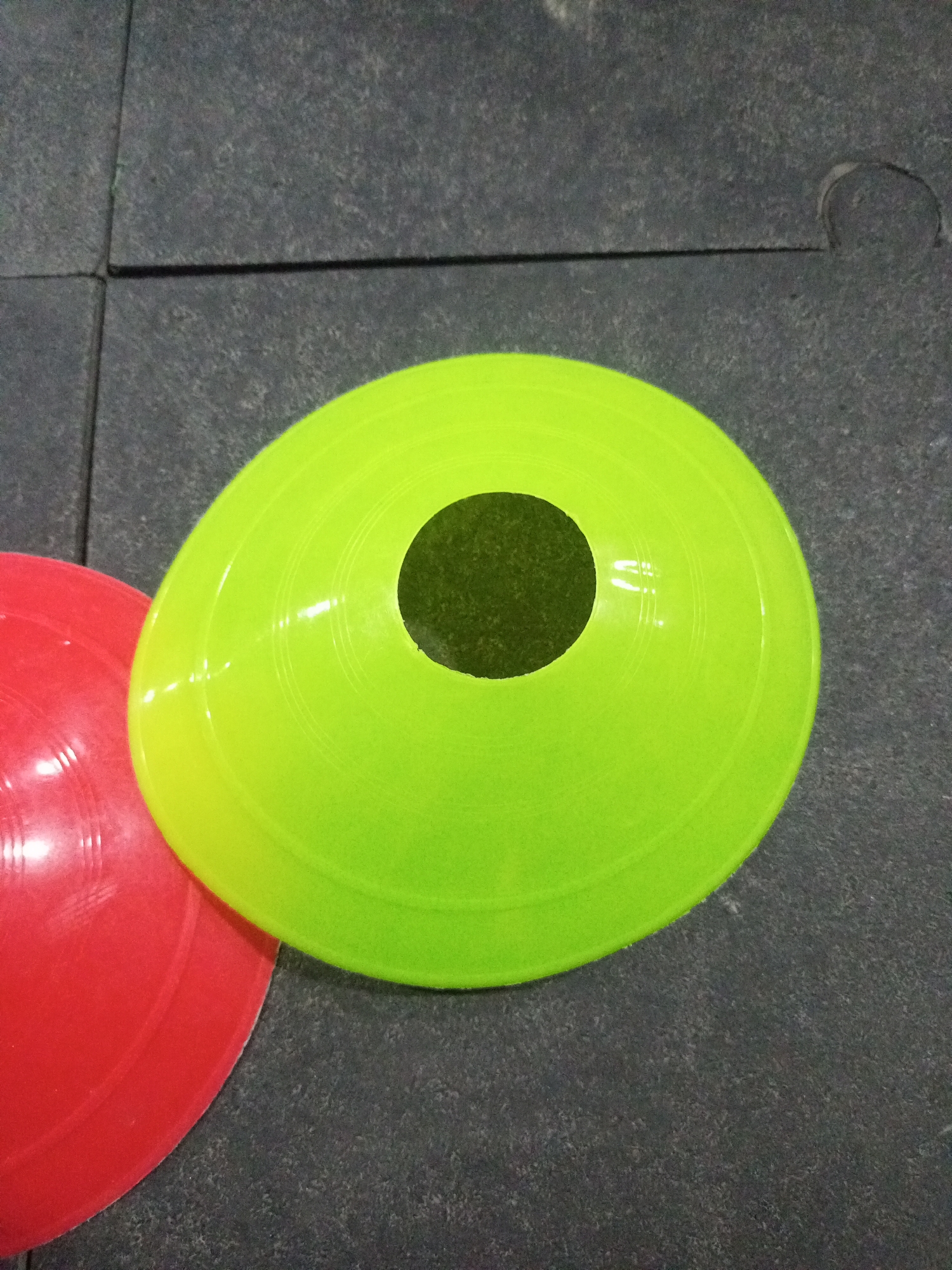 This is a close-up, overhead shot of two brightly colored plastic sports cones on a dark gray, textured surface, likely a gym floor. A large, lime-green cone with a black center hole dominates the right side of the frame. To its left, partially visible, is a smaller red cone. The surface appears to be made of interlocking rubber tiles, with visible seams between them. The lighting casts subtle reflections on the glossy surfaces of the cones. The scene depicts sports training equipment in a utilitarian environment.