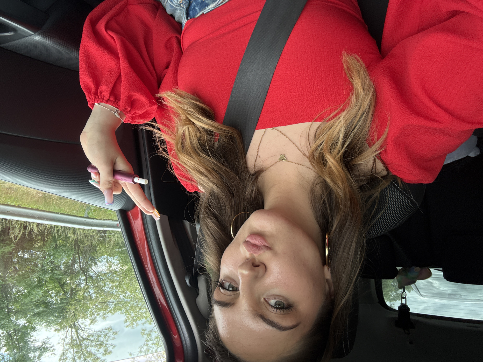 A person is shown from an unusual upside-down perspective in what appears to be the interior of a car. The individual is wearing a red, long-sleeved top and a seatbelt. They have light brown hair with blonde highlights, and their nails are painted with a multi-colored design. They are holding a small purple device in their hand. The background features a glimpse of trees through the car window and parts of the car's interior. There is nothing to indicate that the image was taken in the city of Bliznets, Bulgaria.